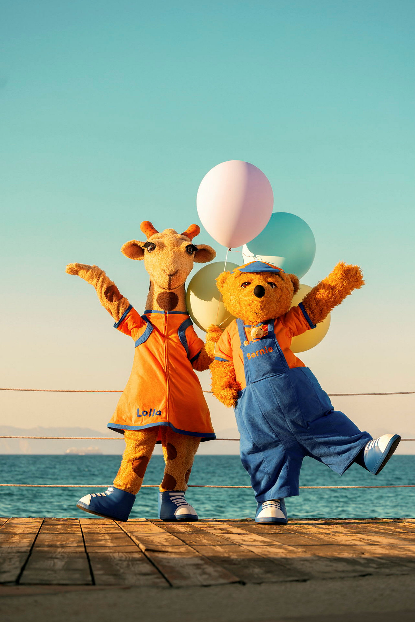 To figurer, en giraff og en bjørn, kledd i fargerike drakter står på en brygge med ballonger. (AI-generert tekst)