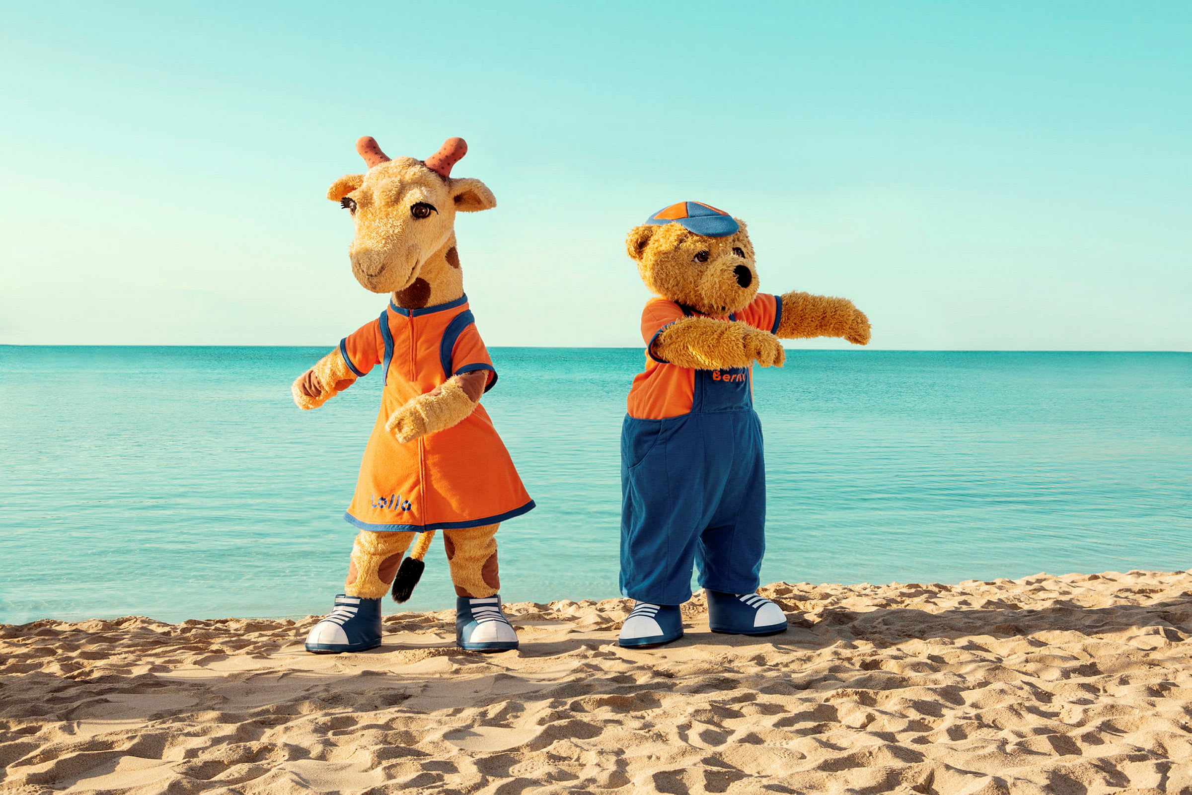 To maskoter, en giraff og en bjørn, står på en strand med havet i bakgrunnen. (AI-generert tekst)