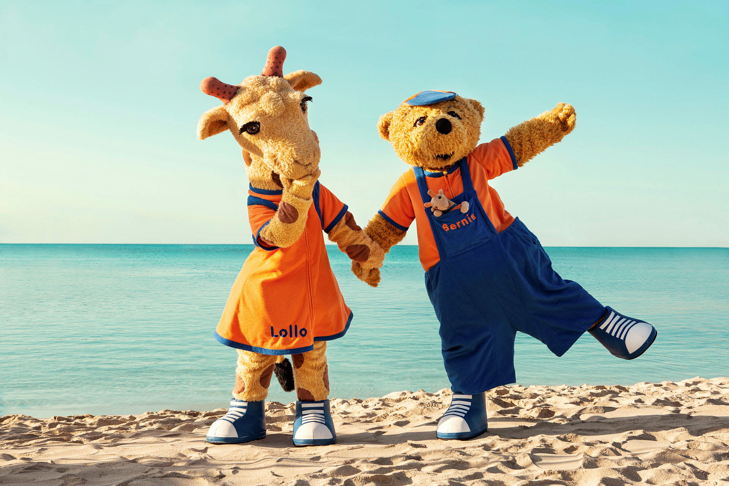 Maskotene Lollo og Bernie står på en sandstrand foran havet. (AI-generert tekst)