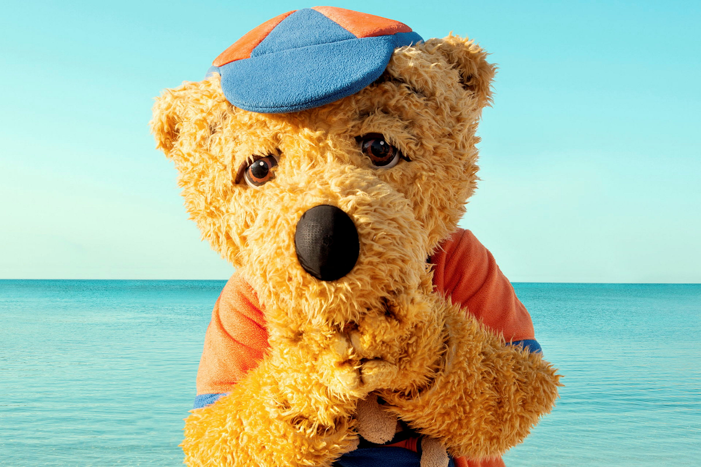 En bamse med caps og klær står ved havet. (AI-generert tekst)