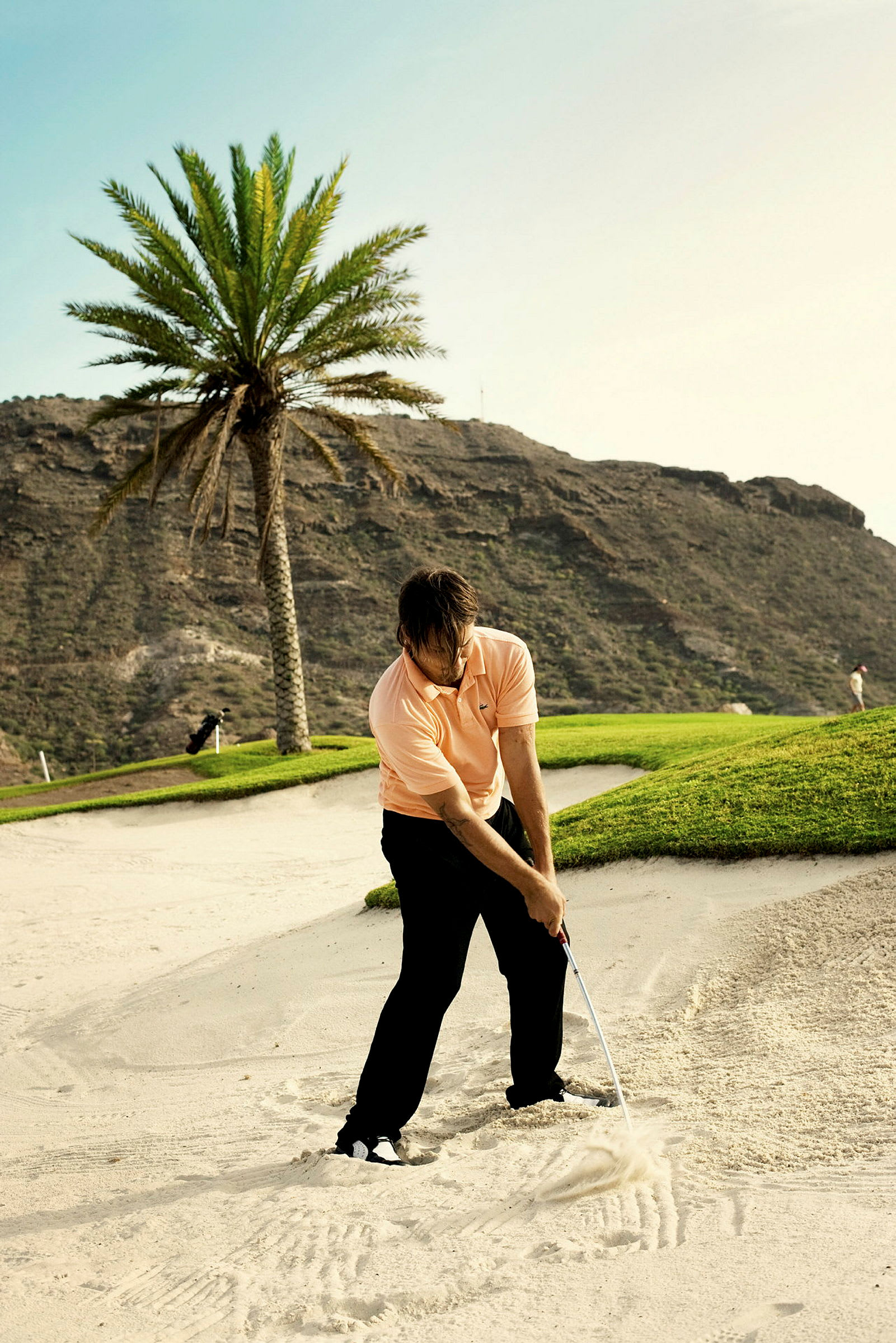 En person spiller golf i en sandbunker med en golfkølle. I bakgrunnen er en palme og fjell. (AI-generert tekst)
