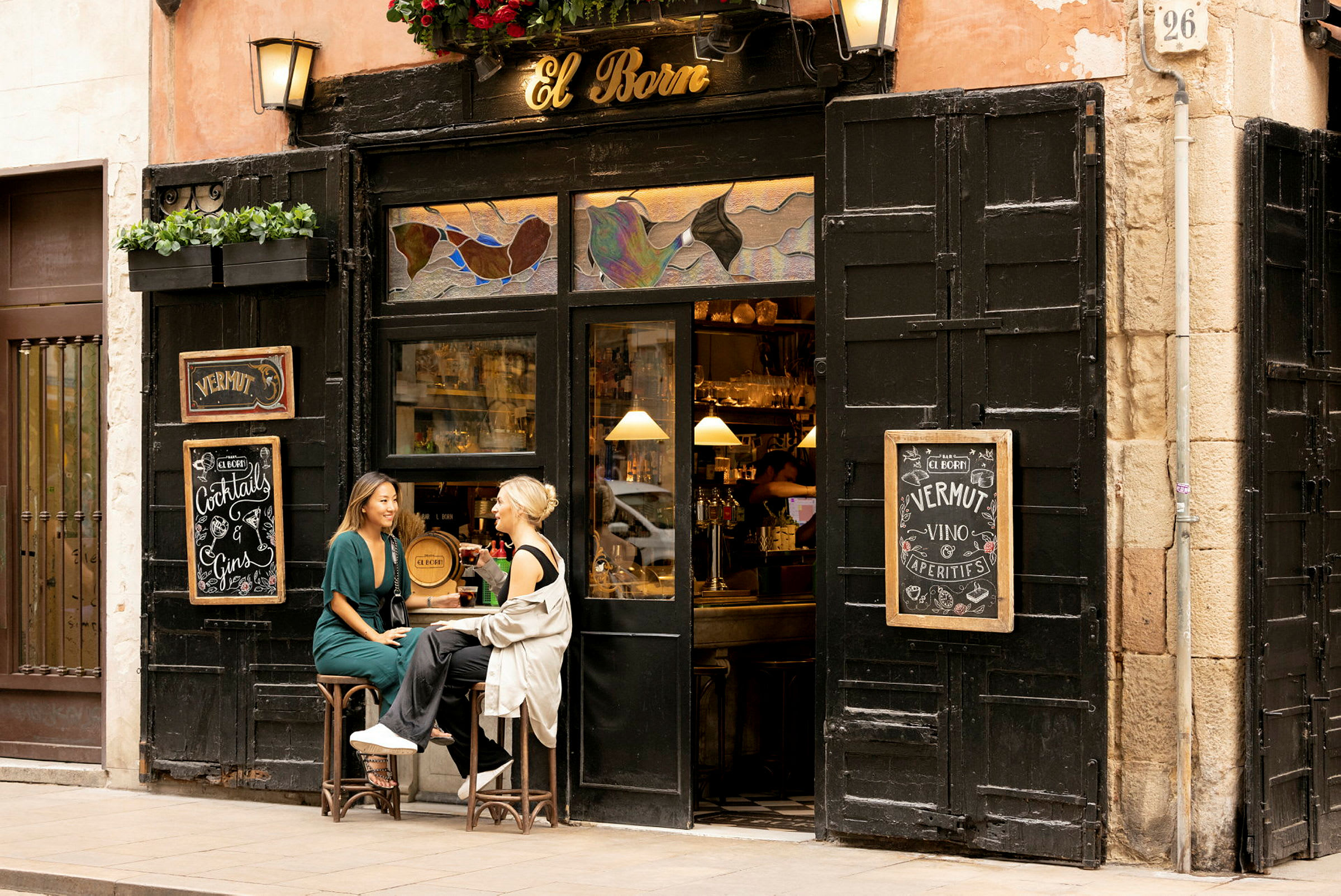 To personer sitter på krakker utenfor en liten bar med skiltet 'El Born'. (AI-generert tekst)