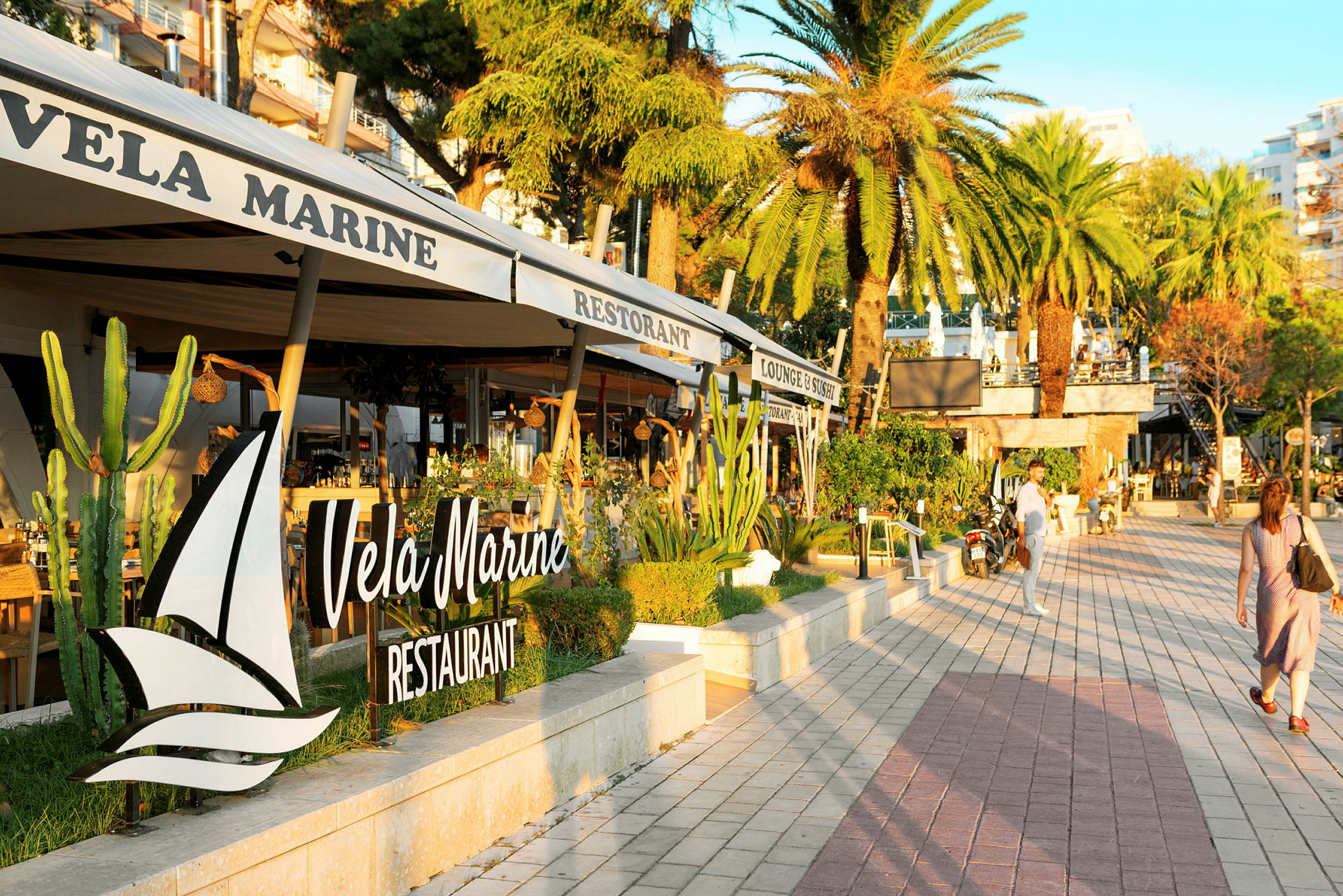 En strandpromenade med palmer og restauranter. Skiltet "Vela Marine Restaurant" er tydelig. (AI-generert tekst)