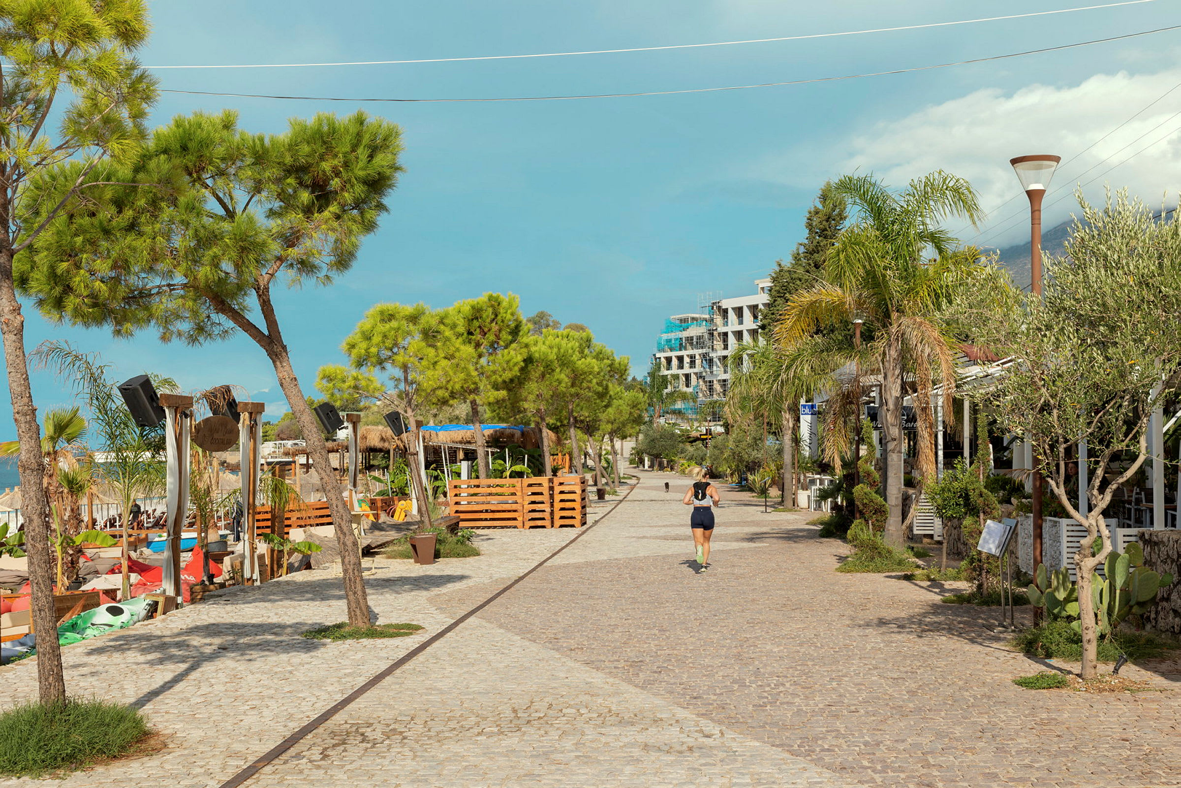 En brosteinslagt promenade med trær, restauranter og et strandområde ved siden av. (AI-generert tekst)