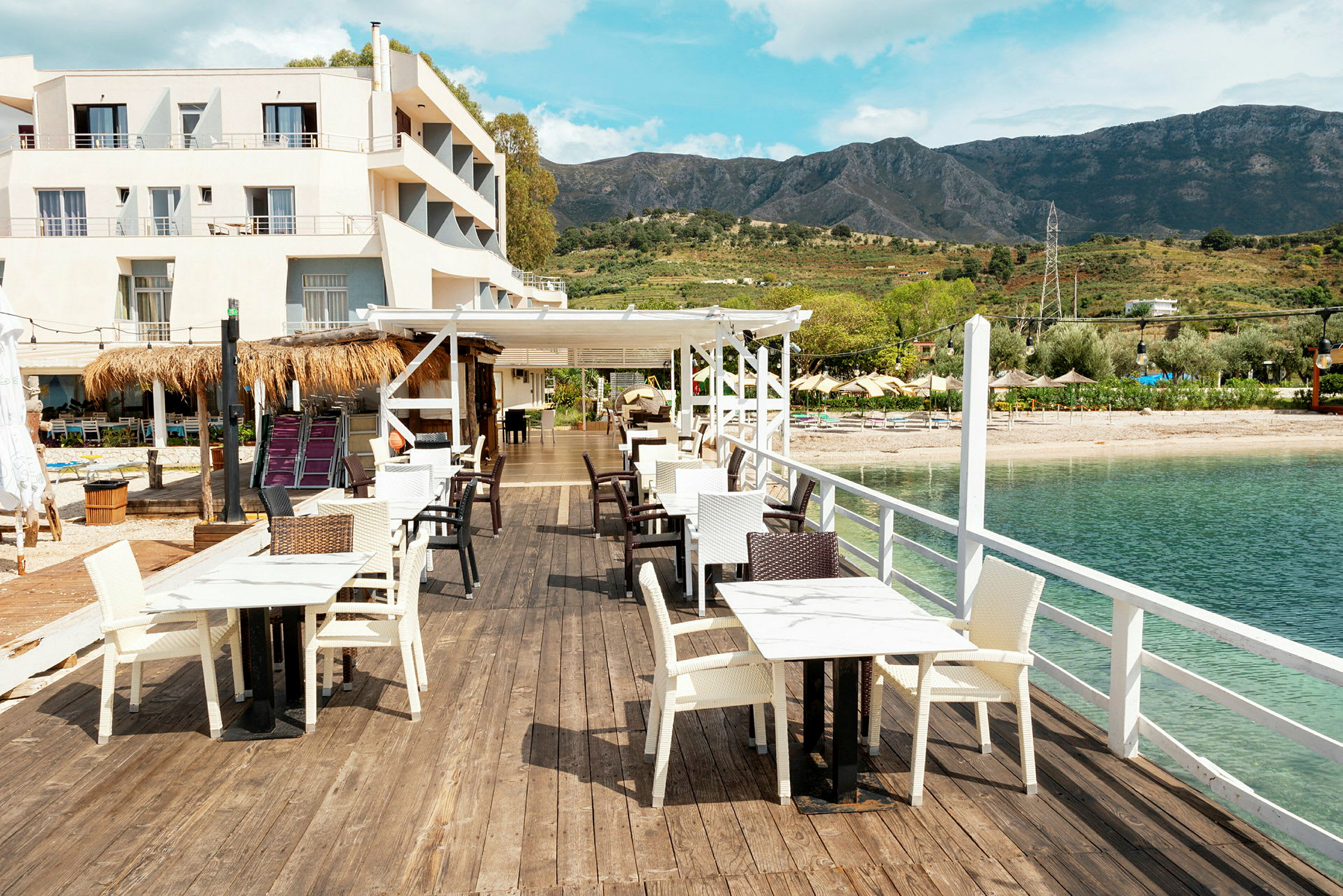 Utendørs serveringsområde med bord og stoler på en trebrygge ved vannet, med et hotell og fjell i bakgrunnen. (AI-generert tekst)