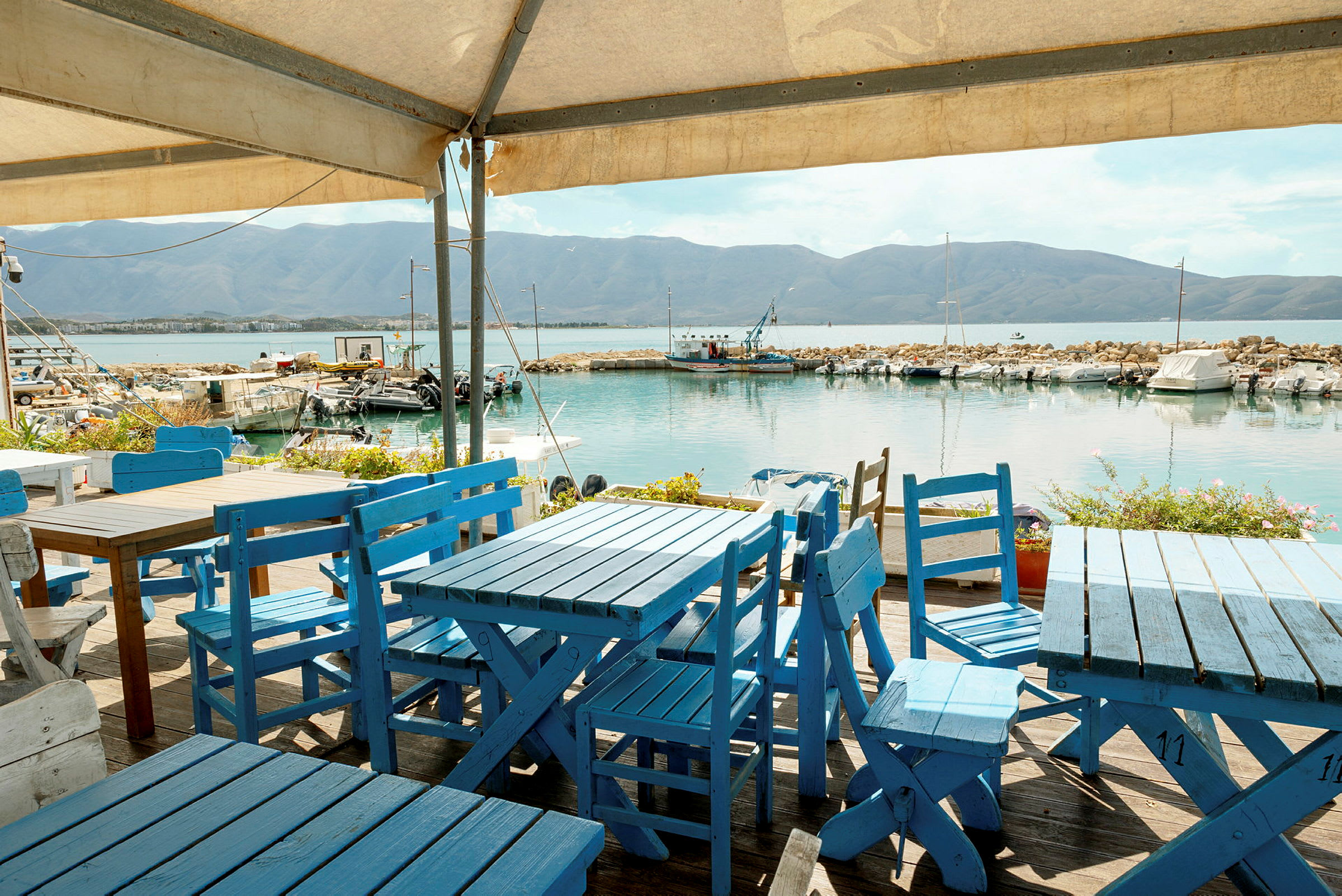 Fire blå trebord med matchende stoler står på en terrasse ved havet. Små båter er fortøyd i vannet foran klipper, og fjell ses i bakgrunnen. (AI-generert tekst)