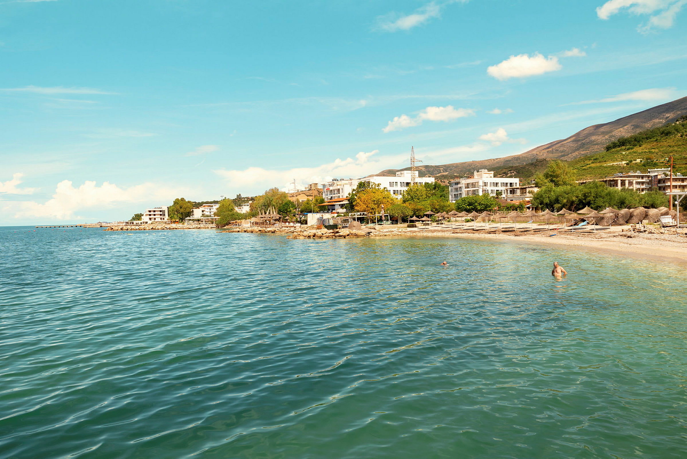 En strand med klart vann og bygninger i bakgrunnen. (AI-generert tekst)