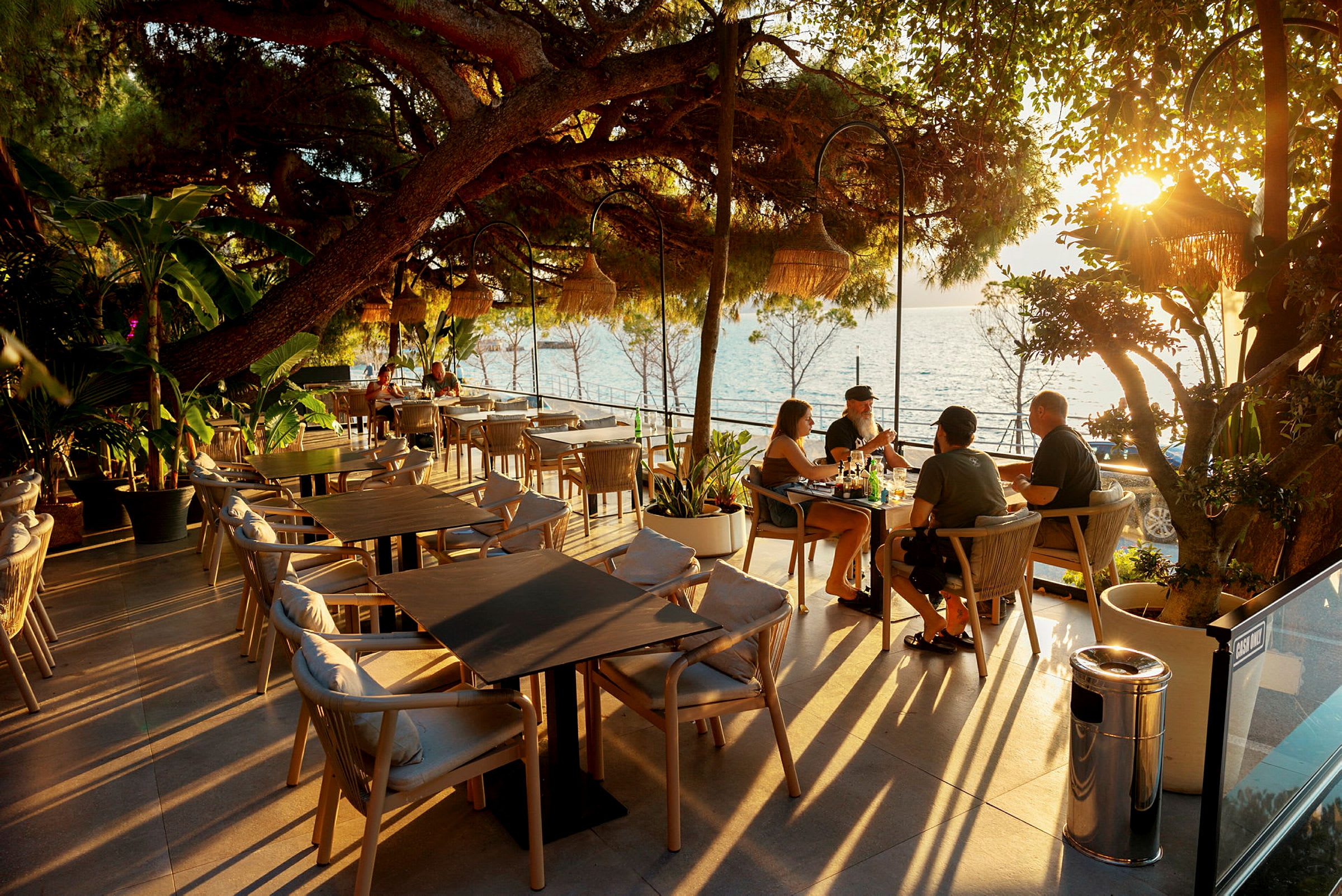 En terrasse med bord og stoler under trær, plassert nær sjøen ved solnedgang. (AI-generert tekst)