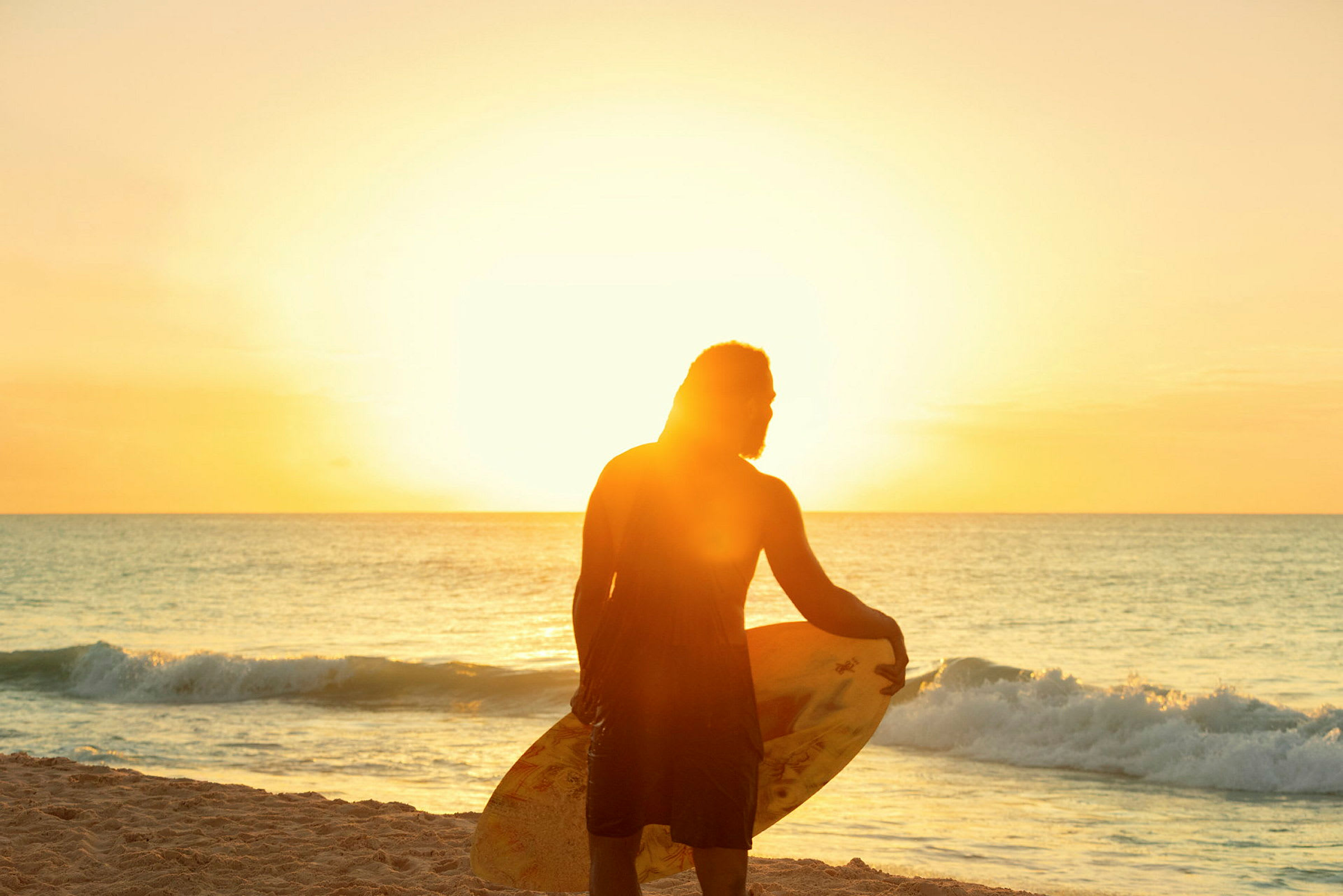 En person står på stranden ved havet, holder et surfebrett og solen lyser sterkt i bakgrunnen. (AI-generert tekst)