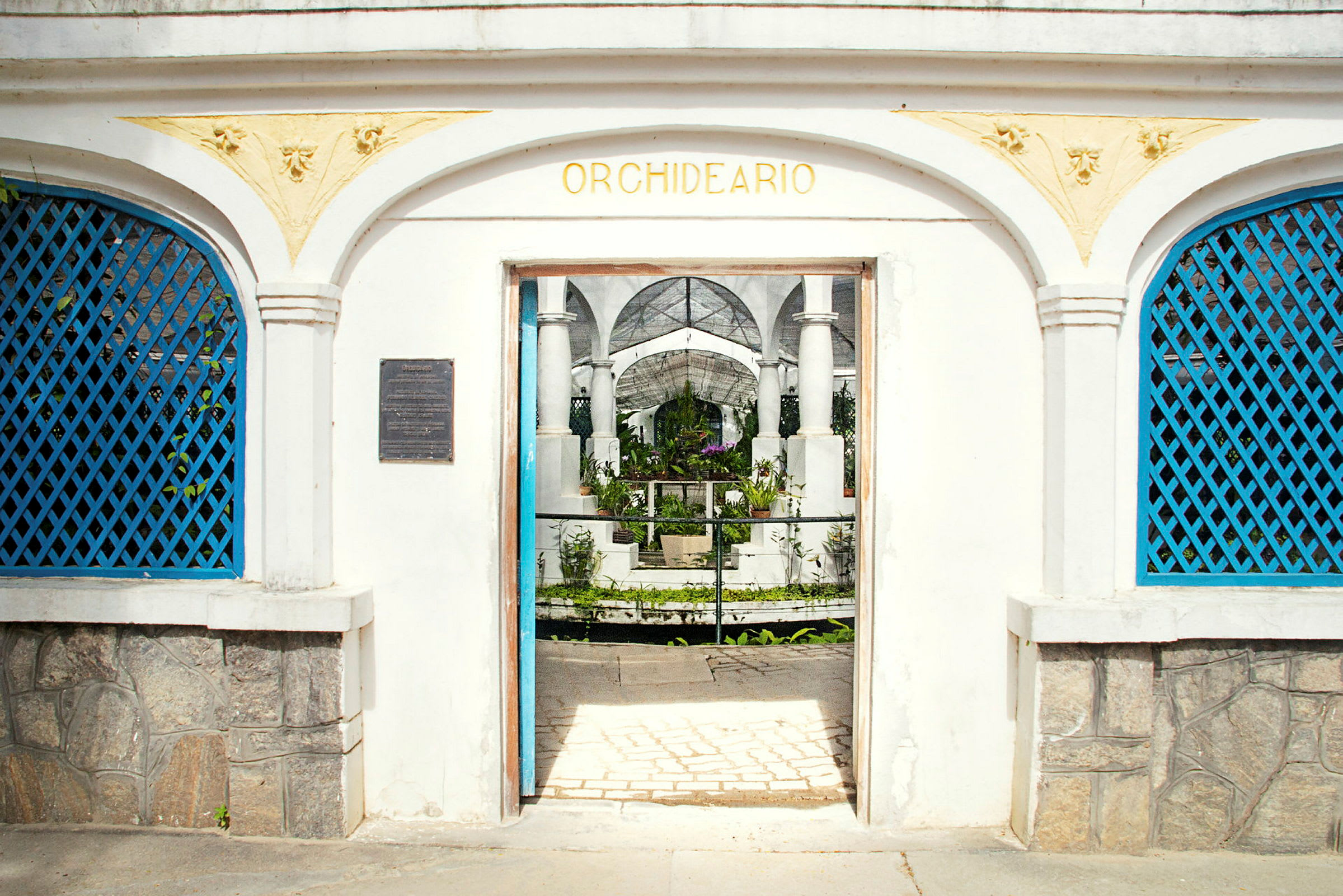 En hvit bygning med en veranda med buer og teksten "Orchideario" over inngangen som fører inn til en hage. (AI-generert tekst)
