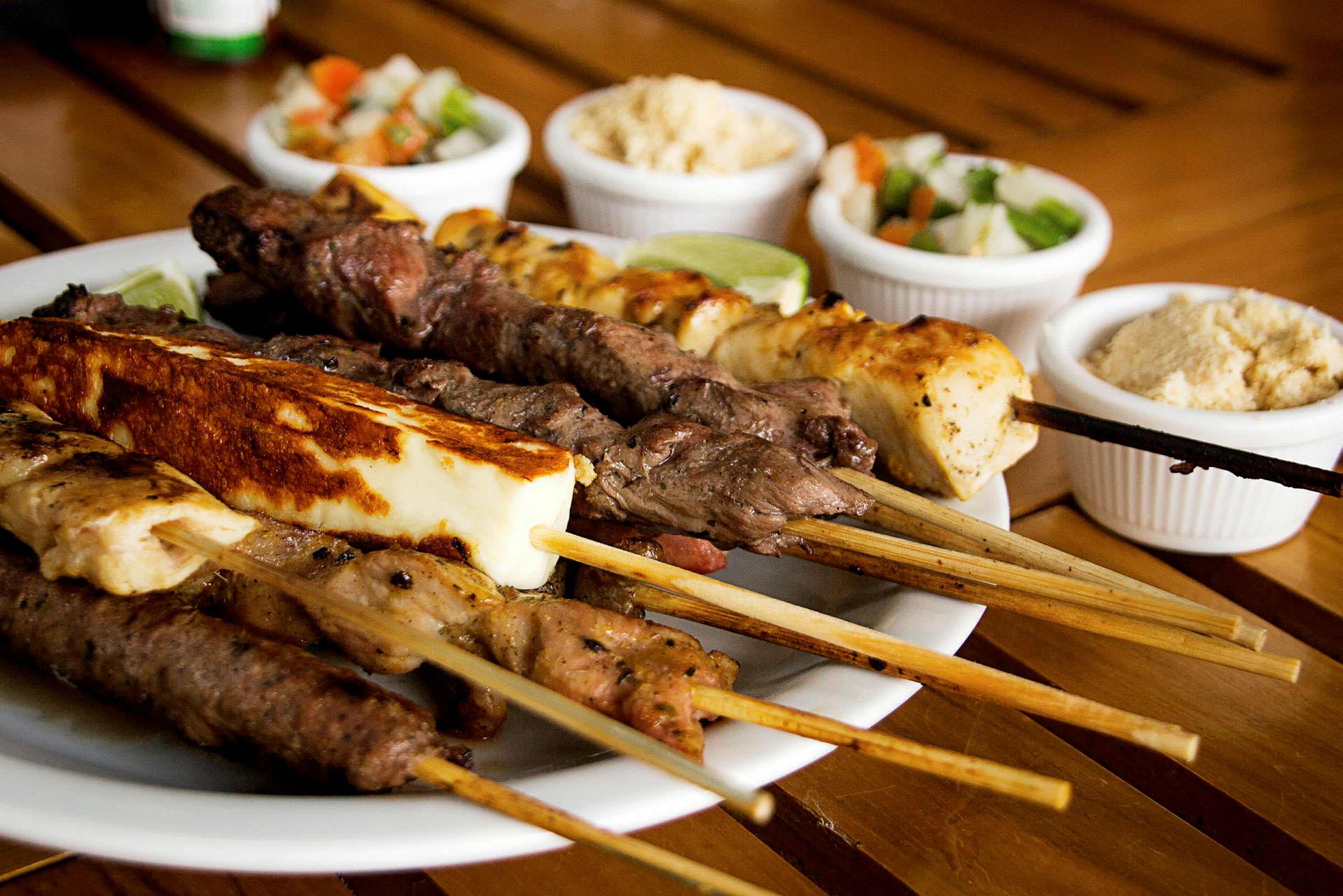 En tallerken med grillspyd av ulike typer kjøtt og ost, med små skåler med tilbehør i bakgrunnen. (AI-generert tekst)