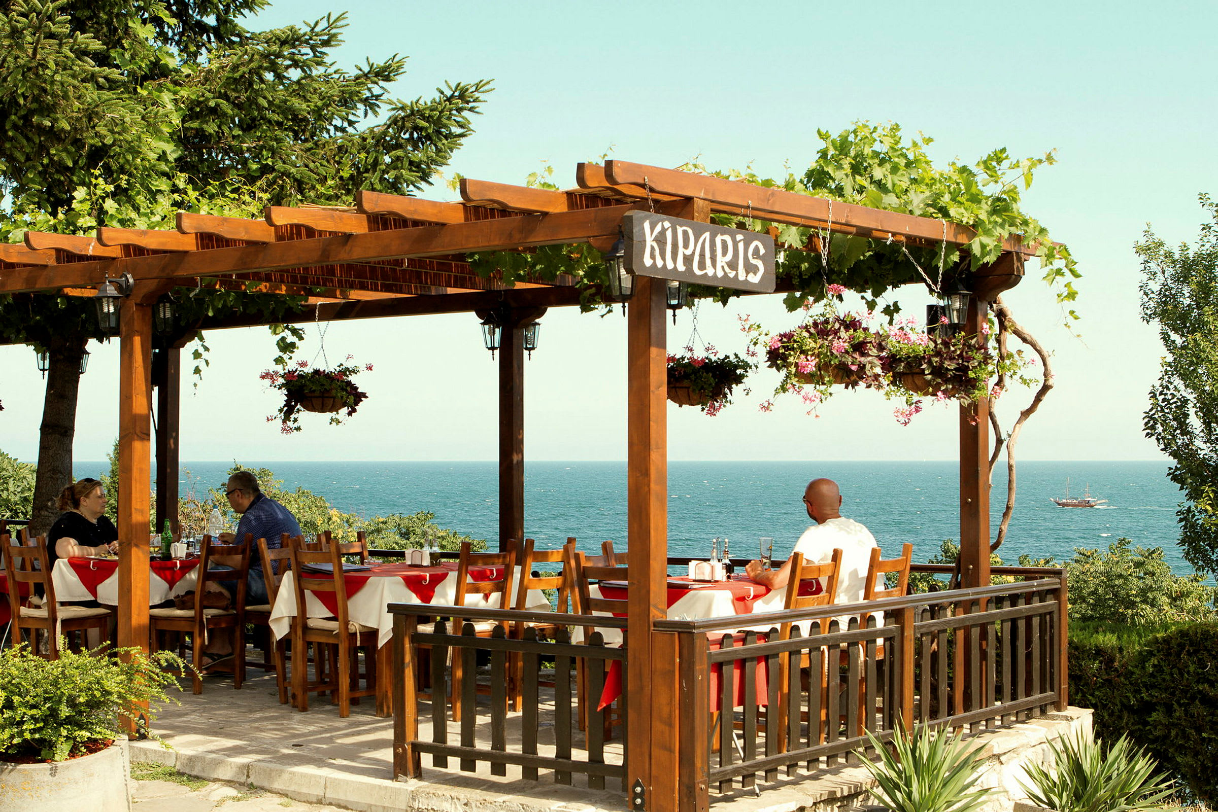 Uteservering med en pergola dekket av planter, havet i bakgrunnen. (AI-generert tekst)