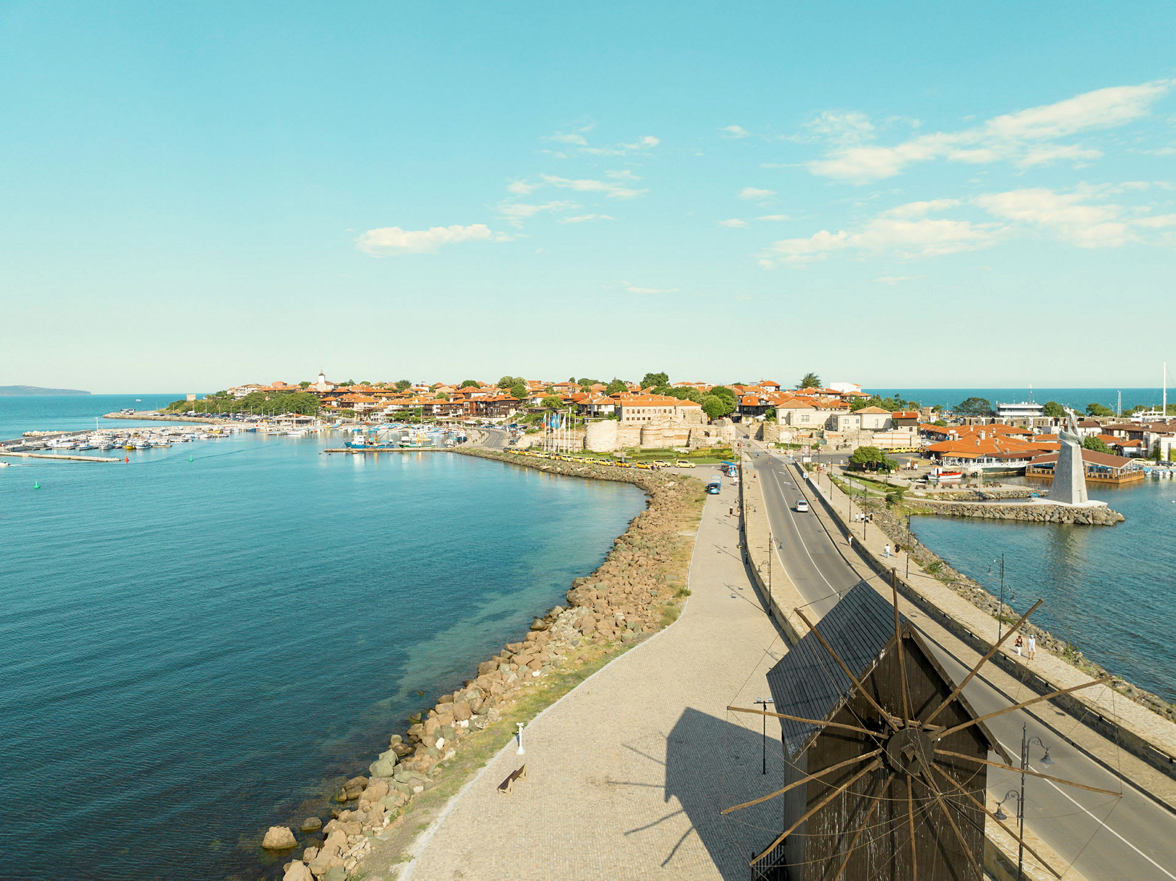 En kystby med bygninger, en vindmølle og en vei i forgrunnen. (AI-generert tekst)