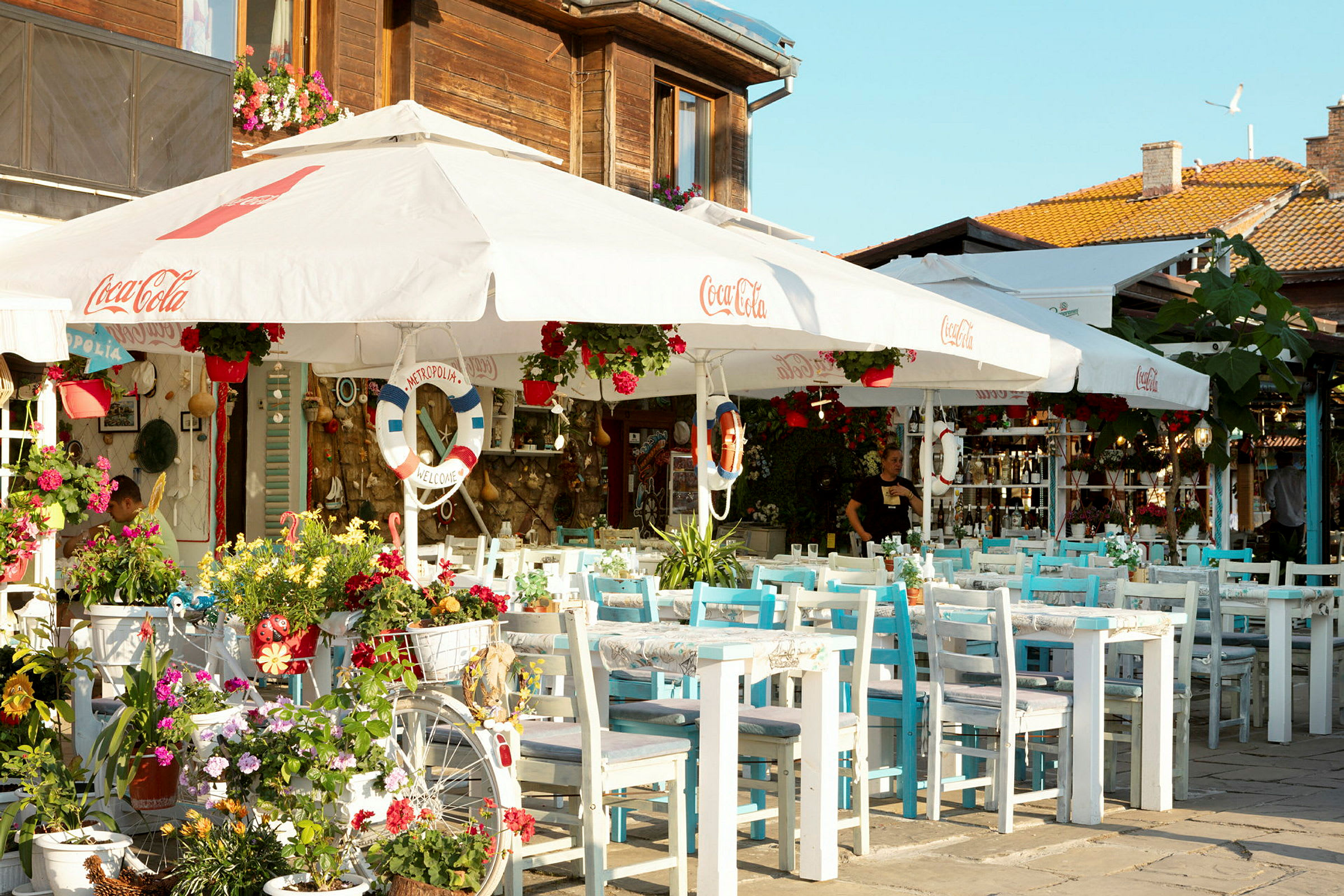 Bildet viser en utecafé med blomster og hvite parasoller. (AI-generert tekst)