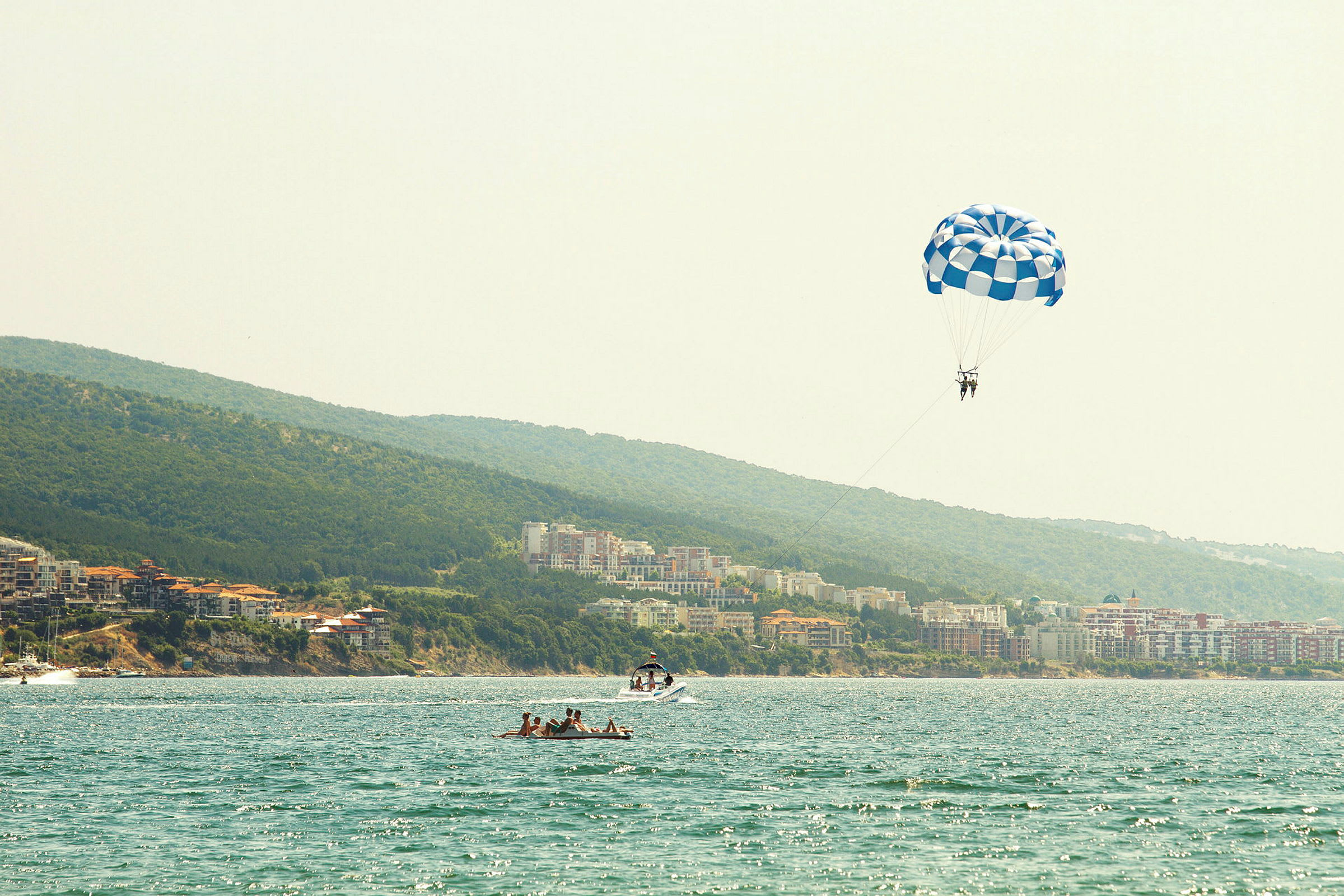 Personer som parasail med en blå og hvit fallskjerm over vannet ved en kyst. (AI-generert tekst)