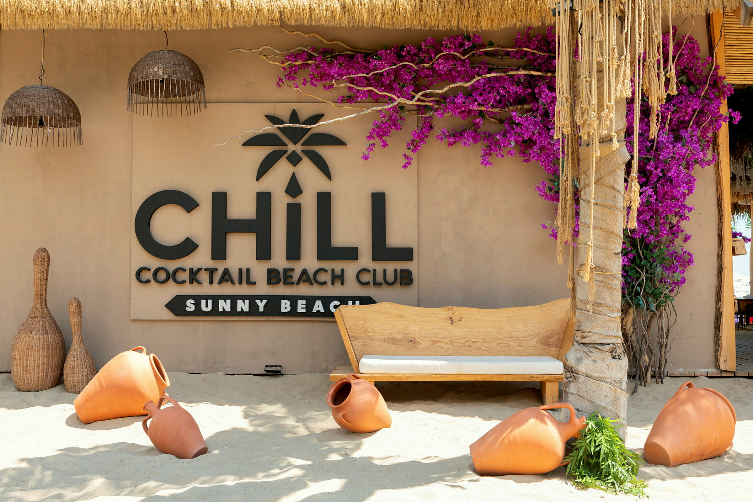 En strandbar med et skilt som sier 'Chill Cocktail Beach Club Sunny Beach', omgitt av terrakottakrukker og fargerike blomster. (AI-generert tekst)