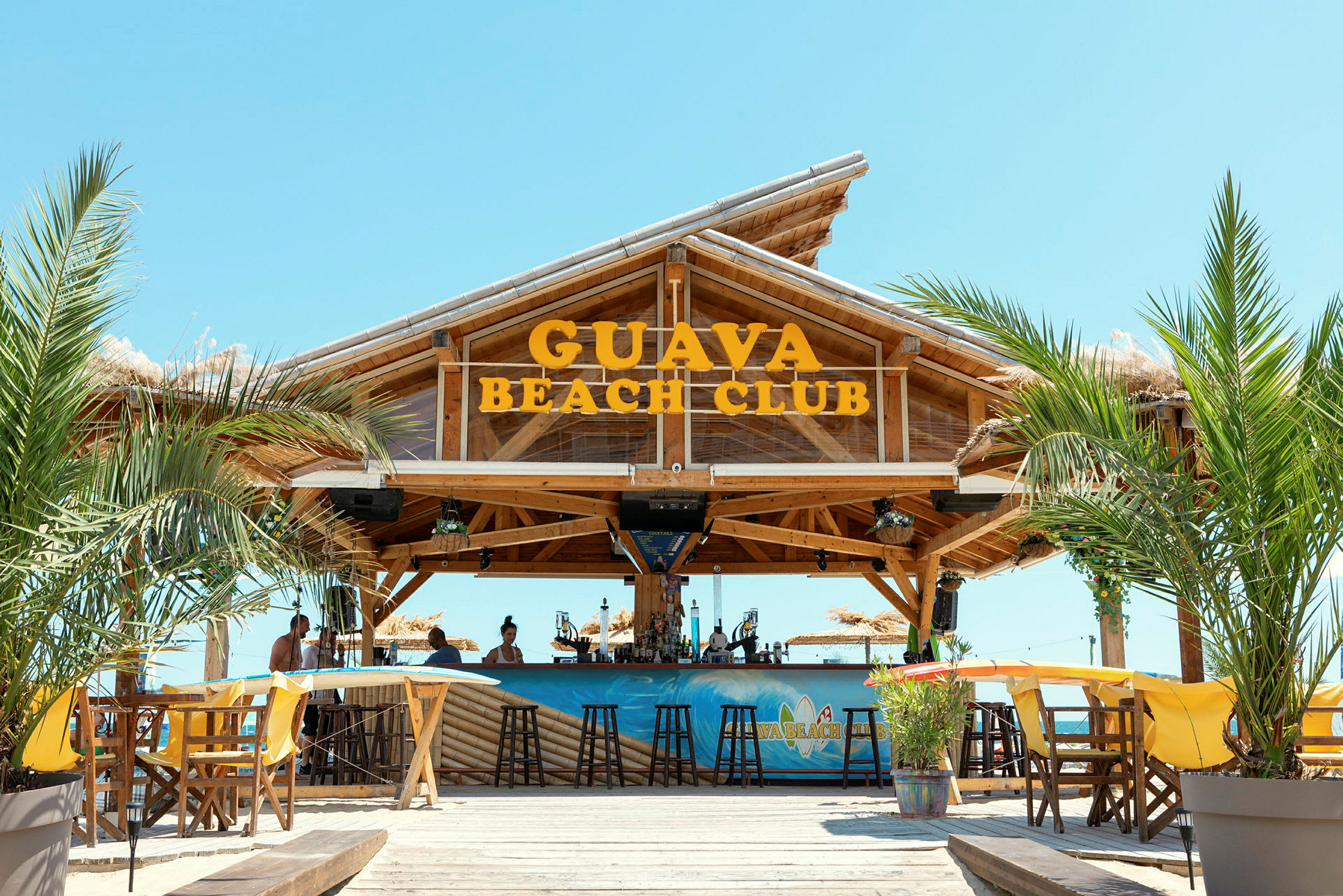 Bilde av Guava Beach Club, en trebar med sitteplasser, palmetrær og klar himmel. (AI-generert tekst)