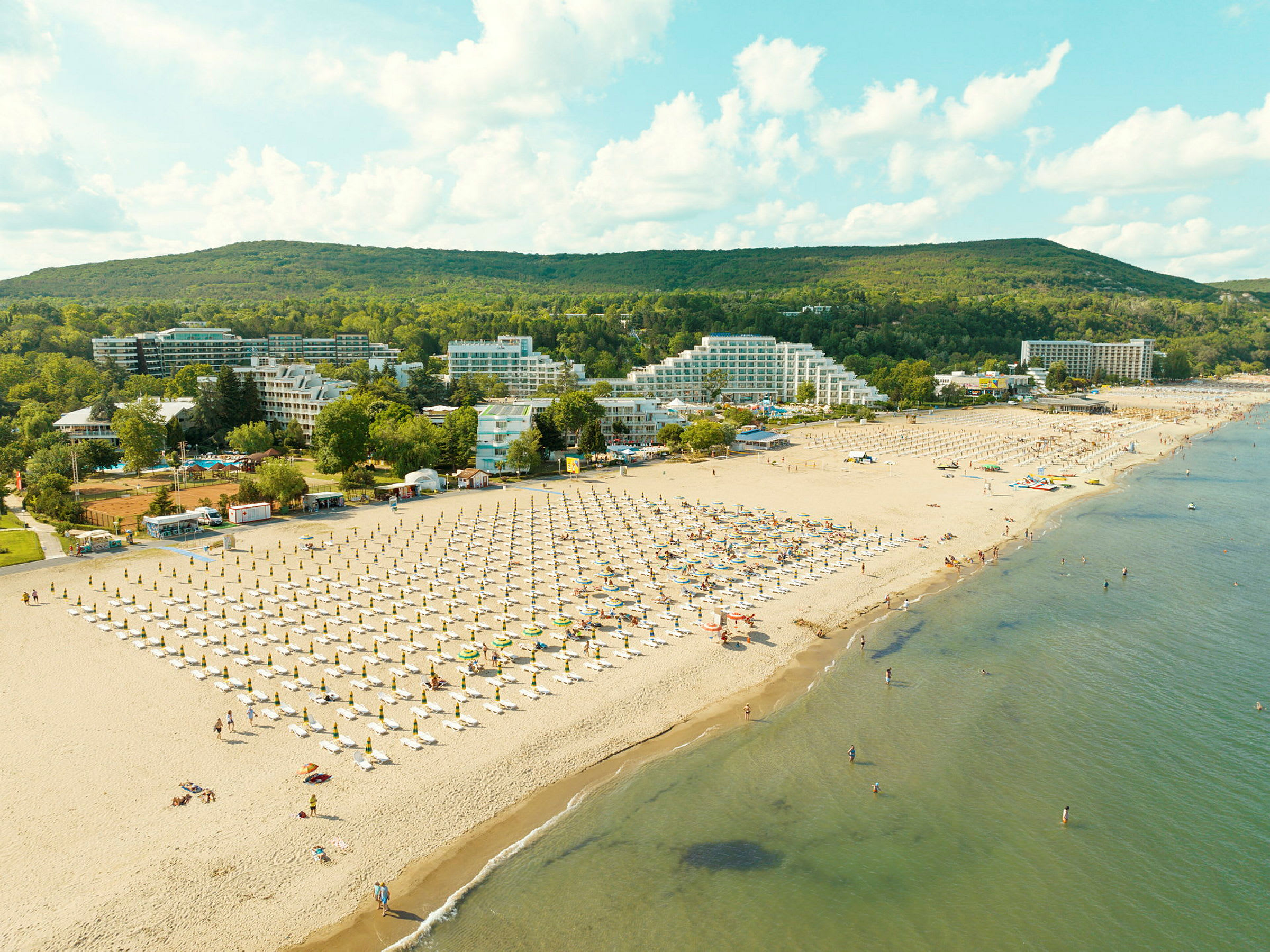 Strand med solsenger og parasoller, omgitt av hotellbygninger og grønne områder. (AI-generert tekst)