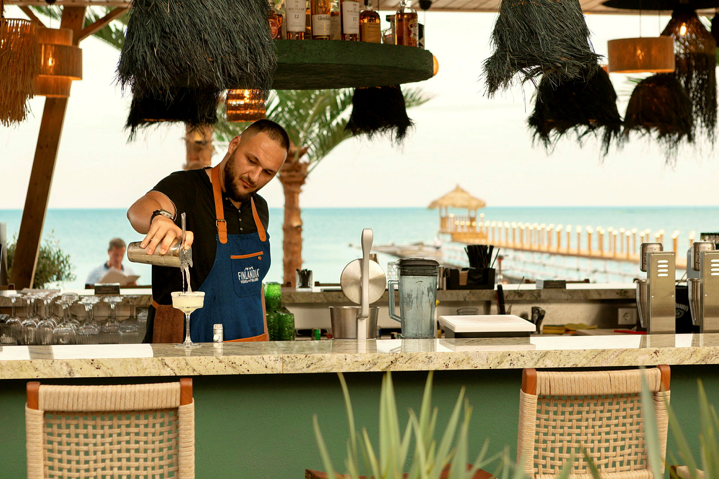 En bartender ved en strandbar heller opp en drink med havet og en brygge i bakgrunnen. (AI-generert tekst)