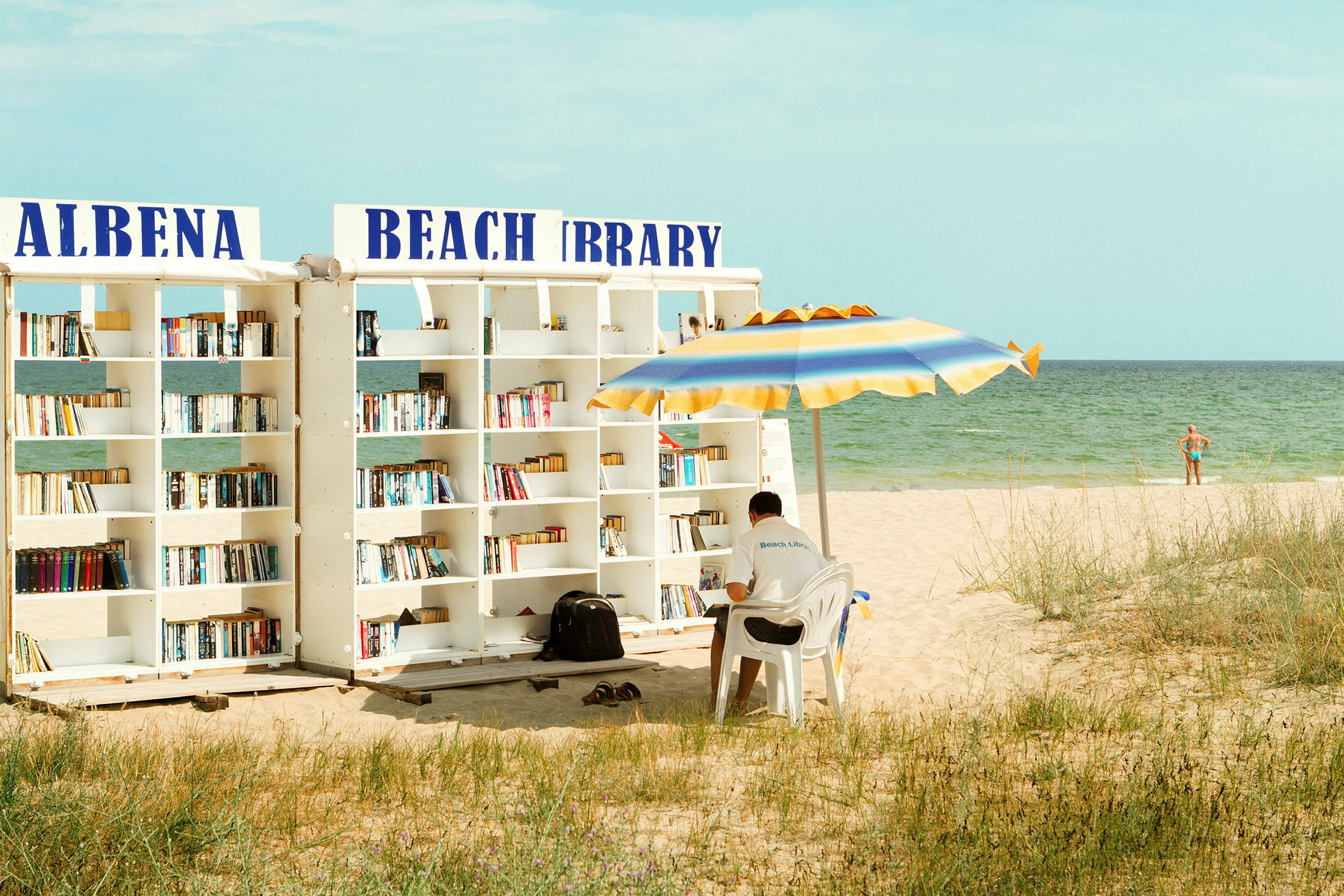 En strand med et utendørs bibliotek og en mann som sitter under en parasoll. (AI-generert tekst)