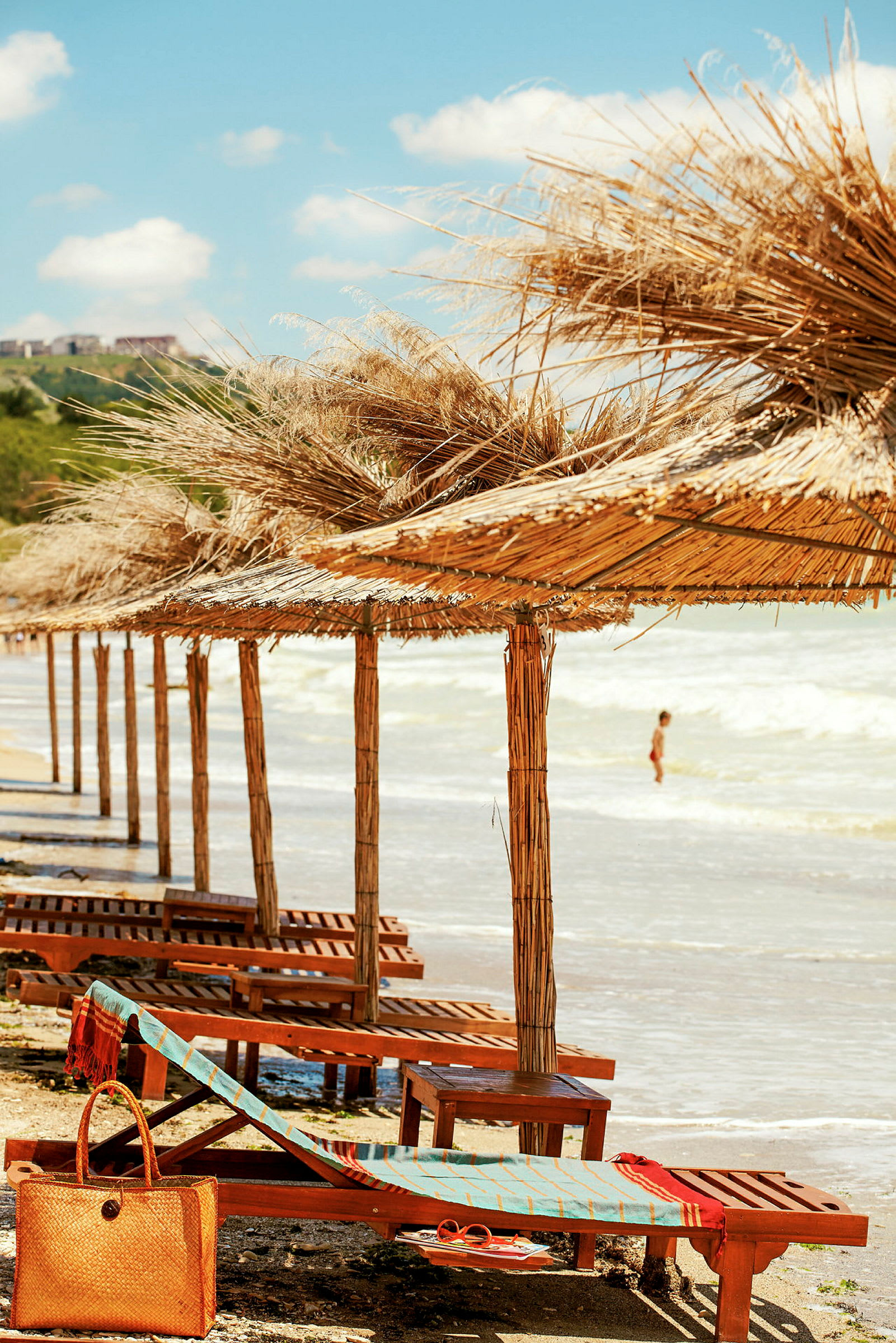 Strand med parasoller av tre og halm samt solsenger, en person er synlig i vannet. (AI-generert tekst)