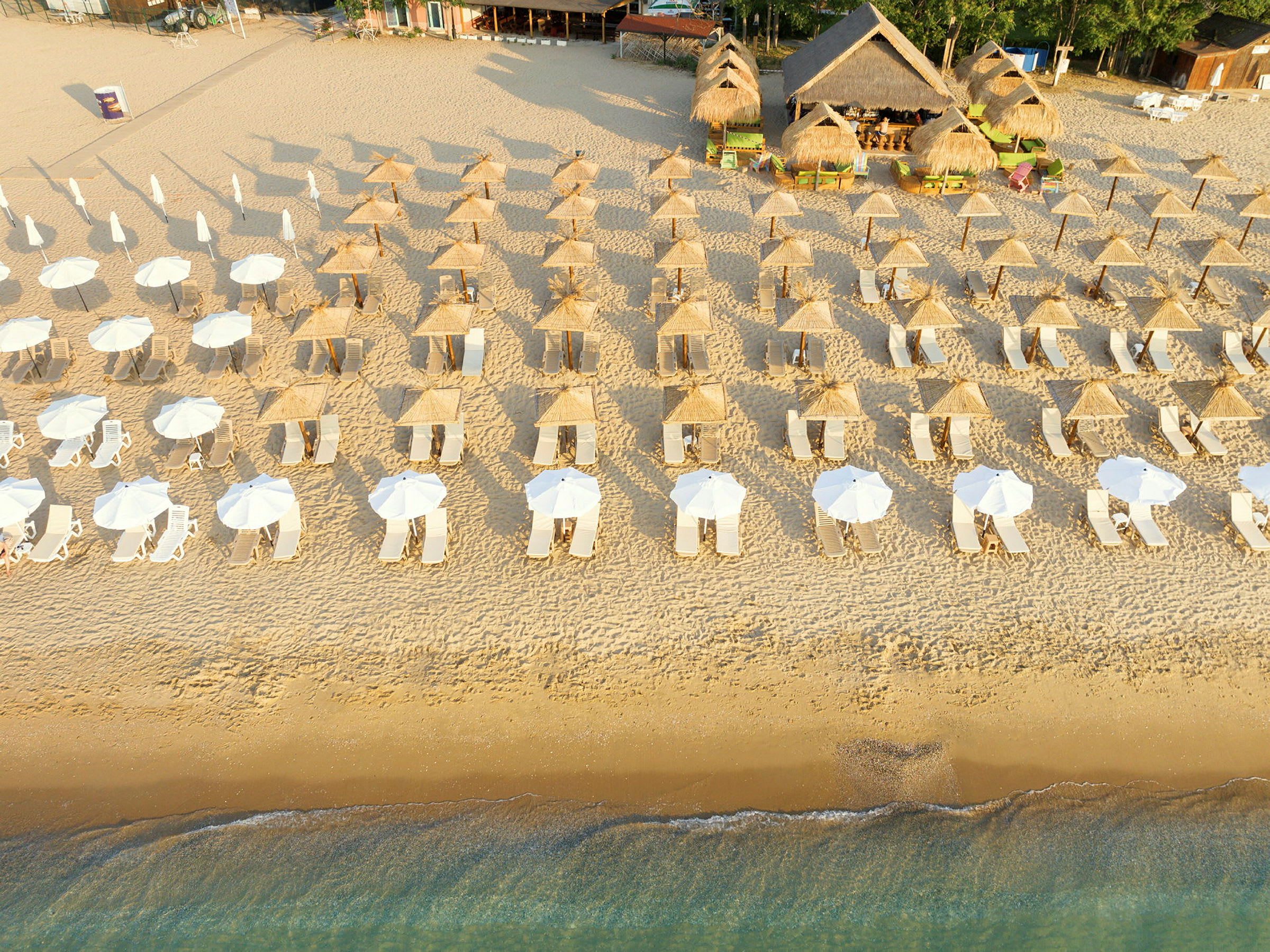 Flyfoto av en strand med rader av solsenger og parasoller, samt strandbar med halmtak i bakgrunnen. (AI-generert tekst)