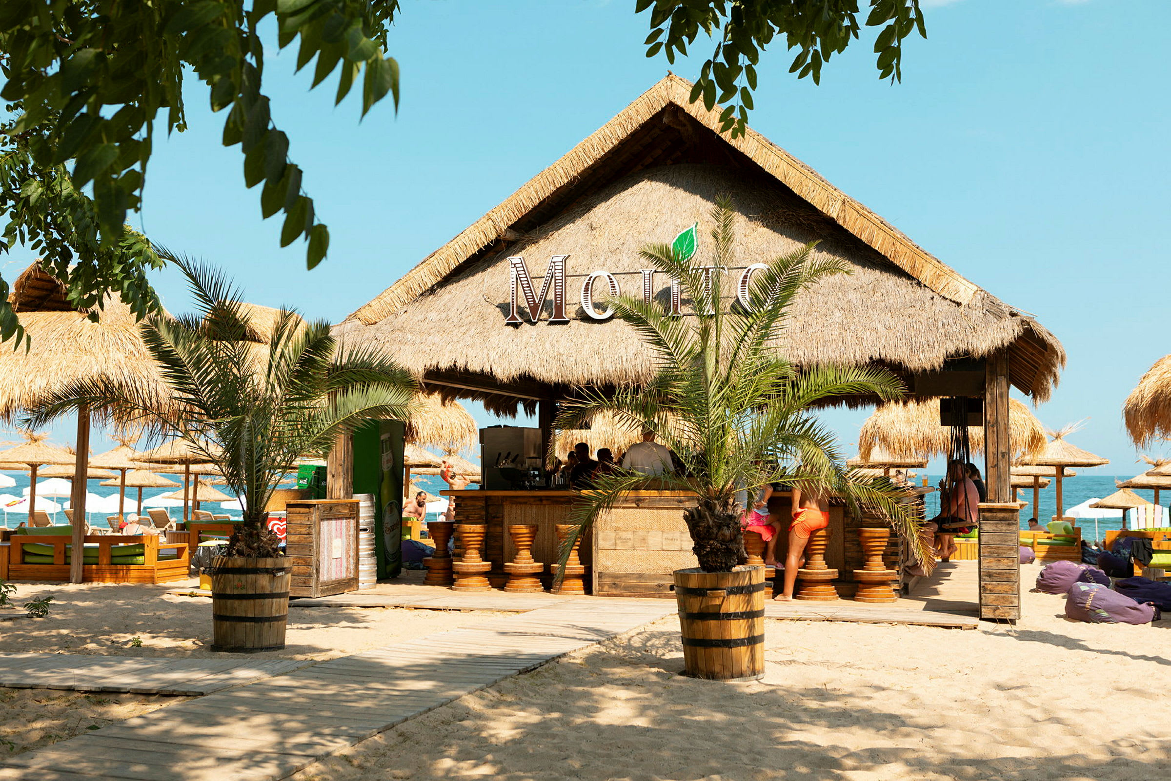 Strandbar med palmebladtak omgitt av sand og parasoller. (AI-generert tekst)