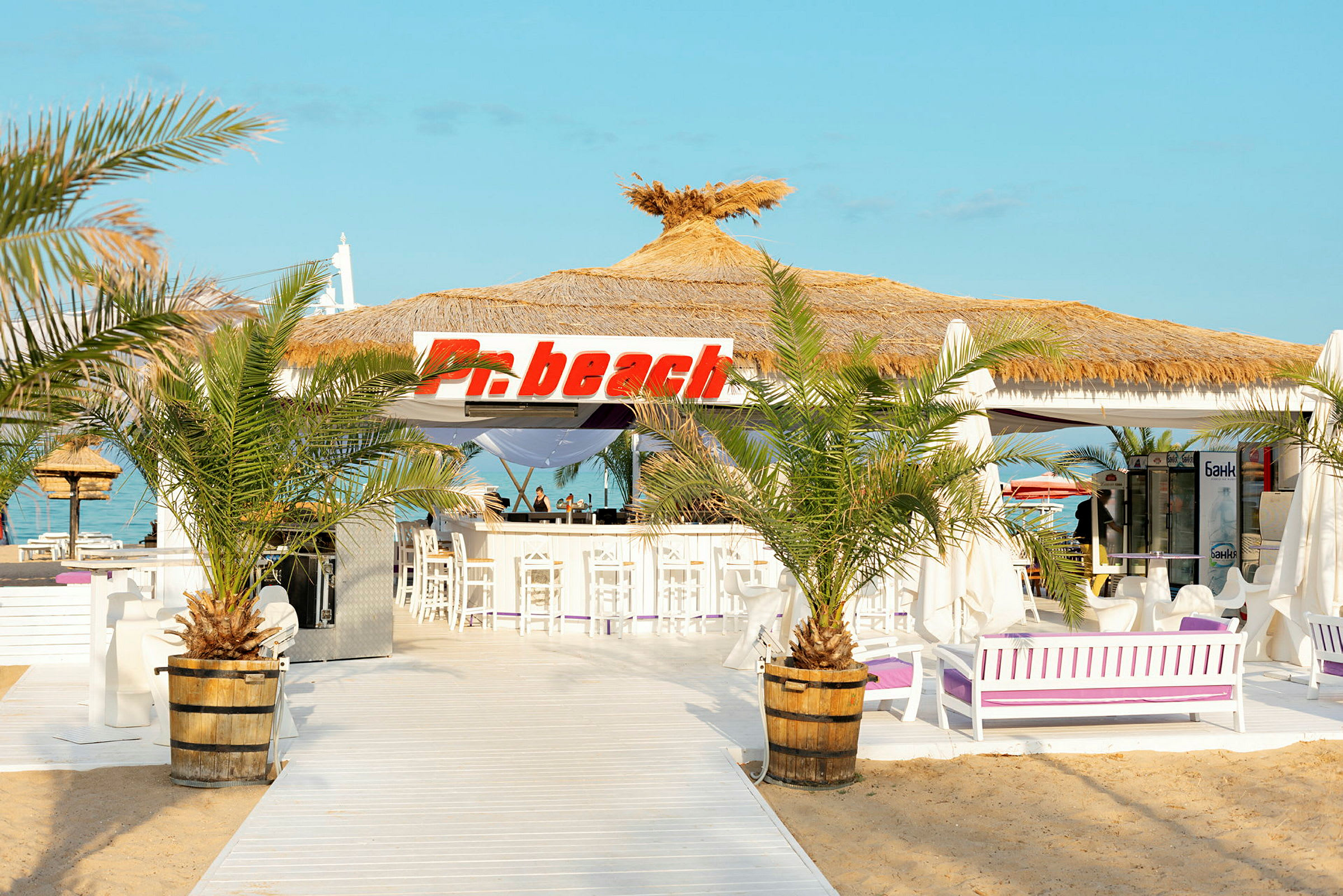 En strandbar med stråtak og skilt som sier 'Pn beach', omgitt av palmer og hvit innredning. (AI-generert tekst)