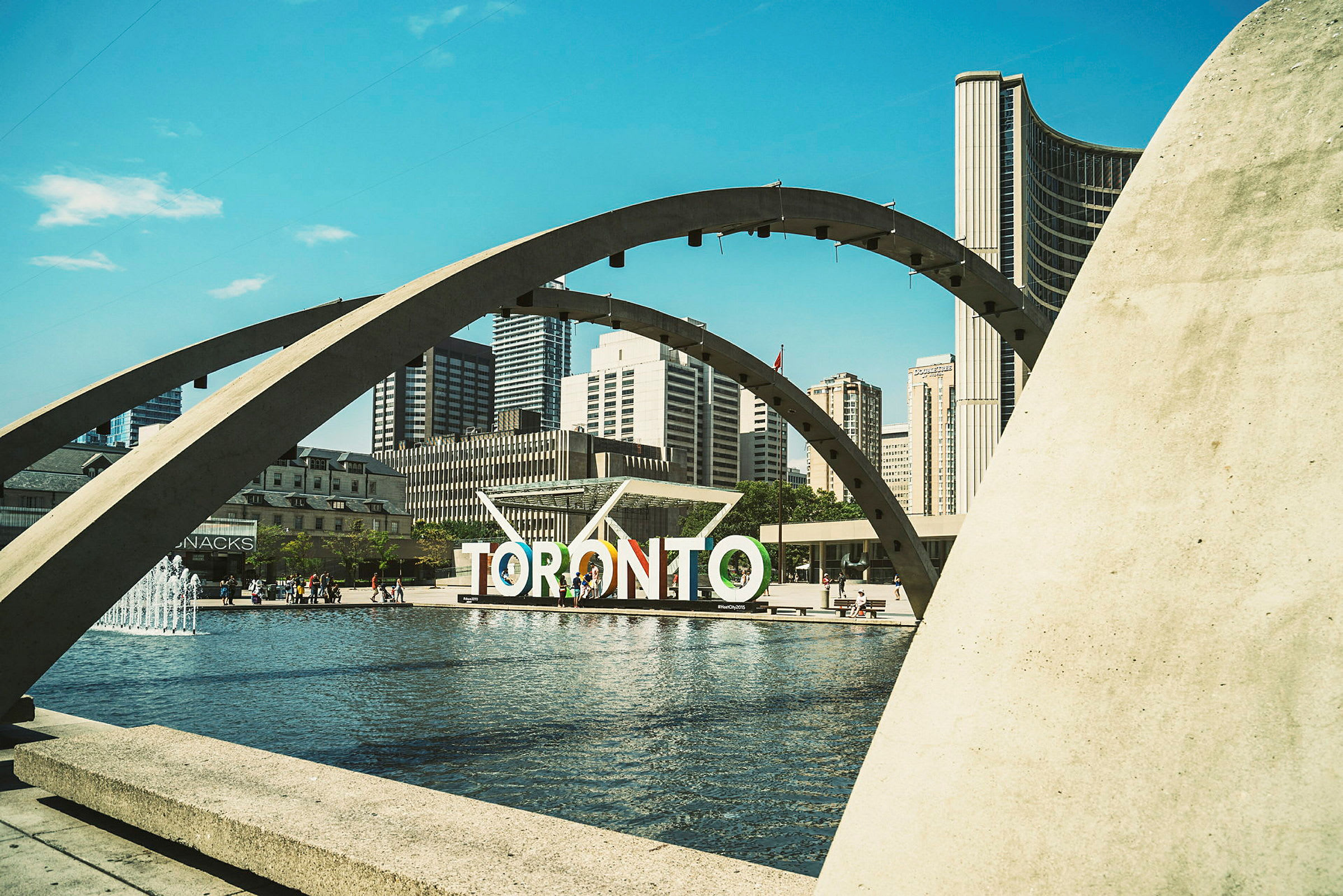 En utsikt over Nathan Phillips Square i Toronto med en fontene, buer og byens skyline i bakgrunnen. (AI-generert tekst)
