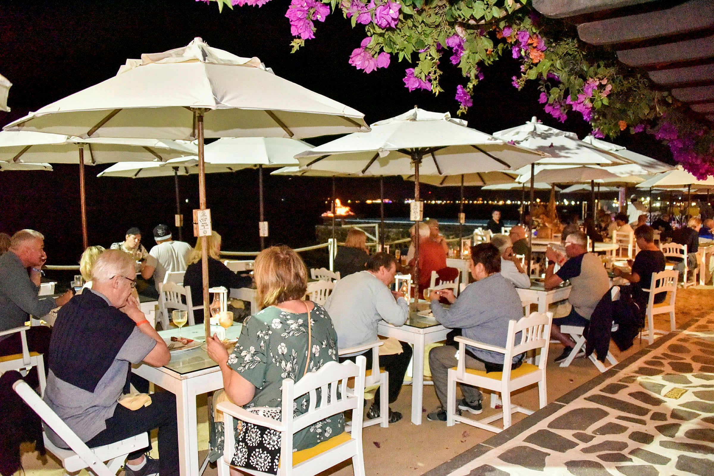 Folk sitter ved utendørsbord under hvite parasoller på en kveldsrestaurant, med blomstrende planter i forgrunnen og havet i bakgrunnen. (AI-generert tekst)