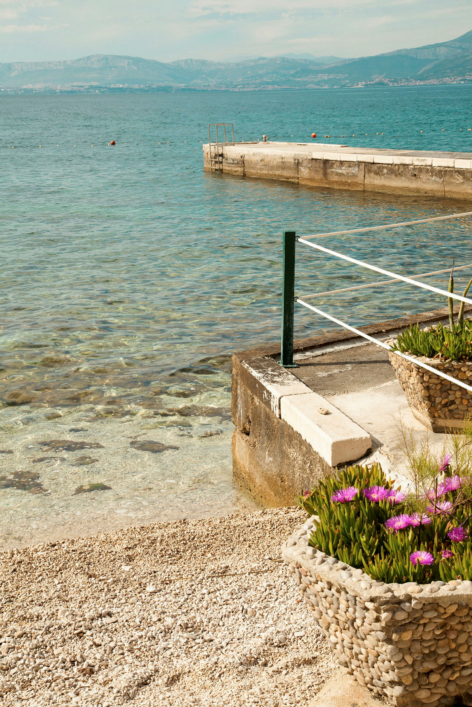 Strand med stein, blomsterpotte med lilla blomster og brygge ved klart vann. (AI-generert tekst)