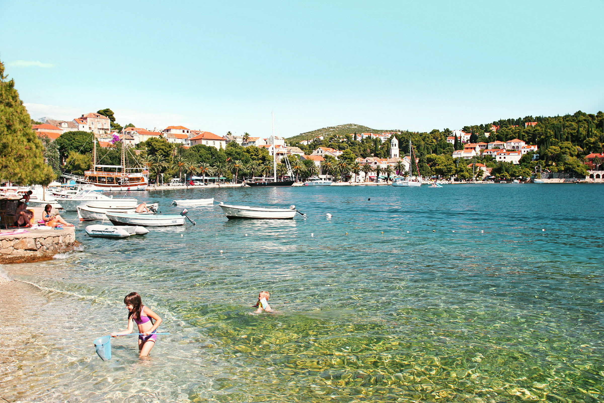 Strand med klart vann, småbåter og en by i bakgrunnen. (AI-generert tekst)