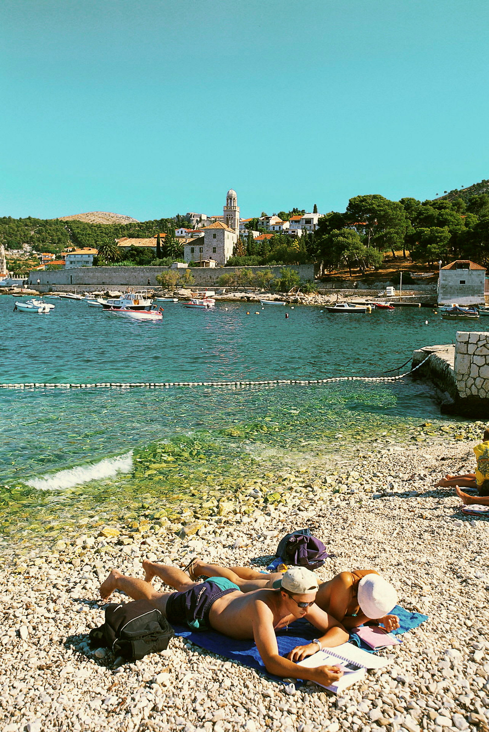 Personer som ligger på en steinete strand ved sjøen med solfylte bygninger i bakgrunnen. (AI-generert tekst)