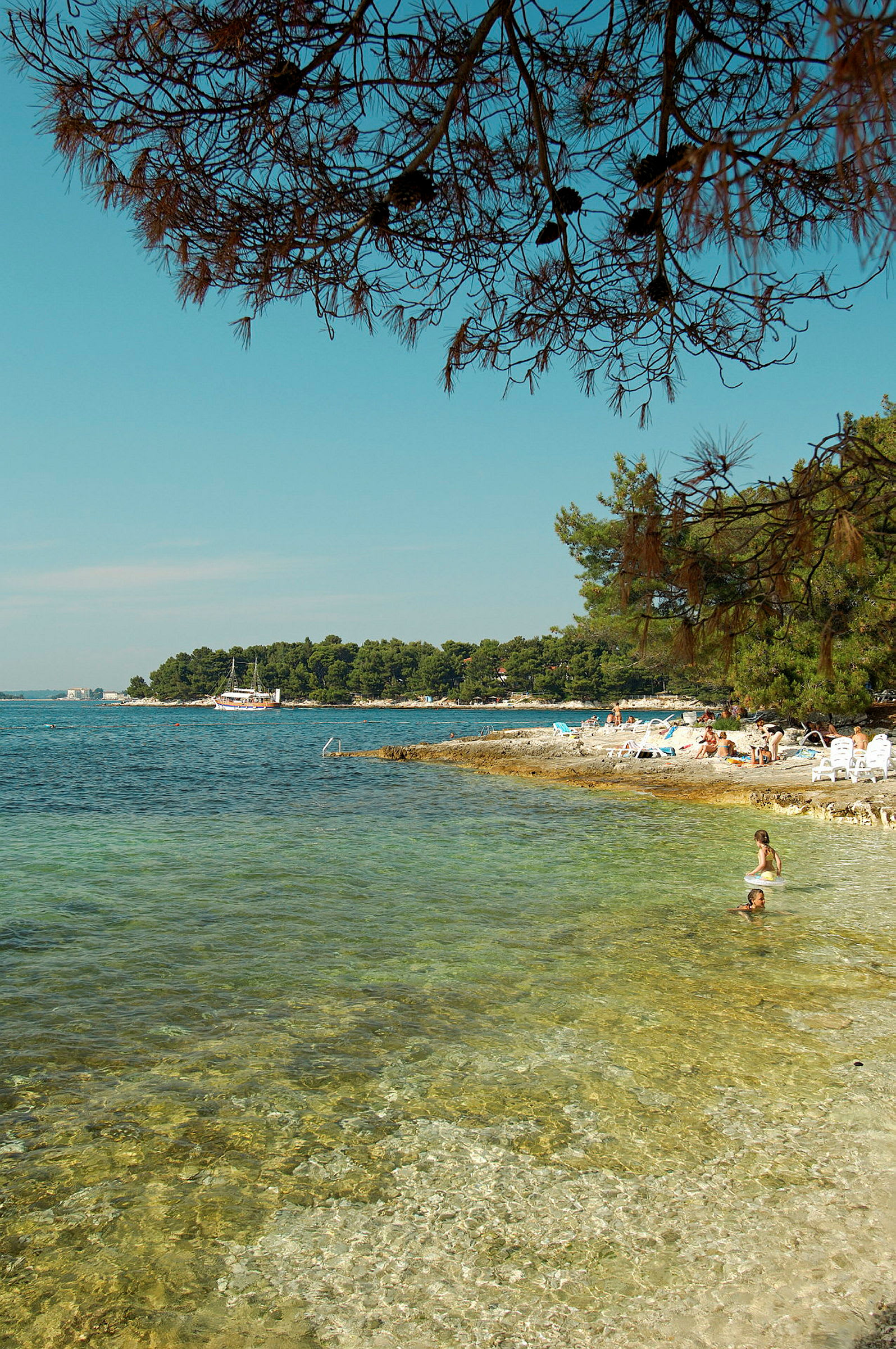 Strand med klart vann og folk som bader, omgitt av trær og en båt lenger unna. (AI-generert tekst)