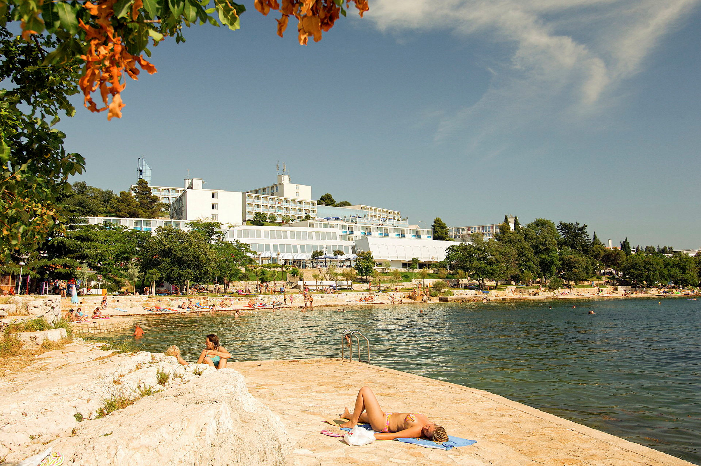 En person ligger på et håndkle ved vannet med hotellbygninger og en strand i bakgrunnen. (AI-generert tekst)