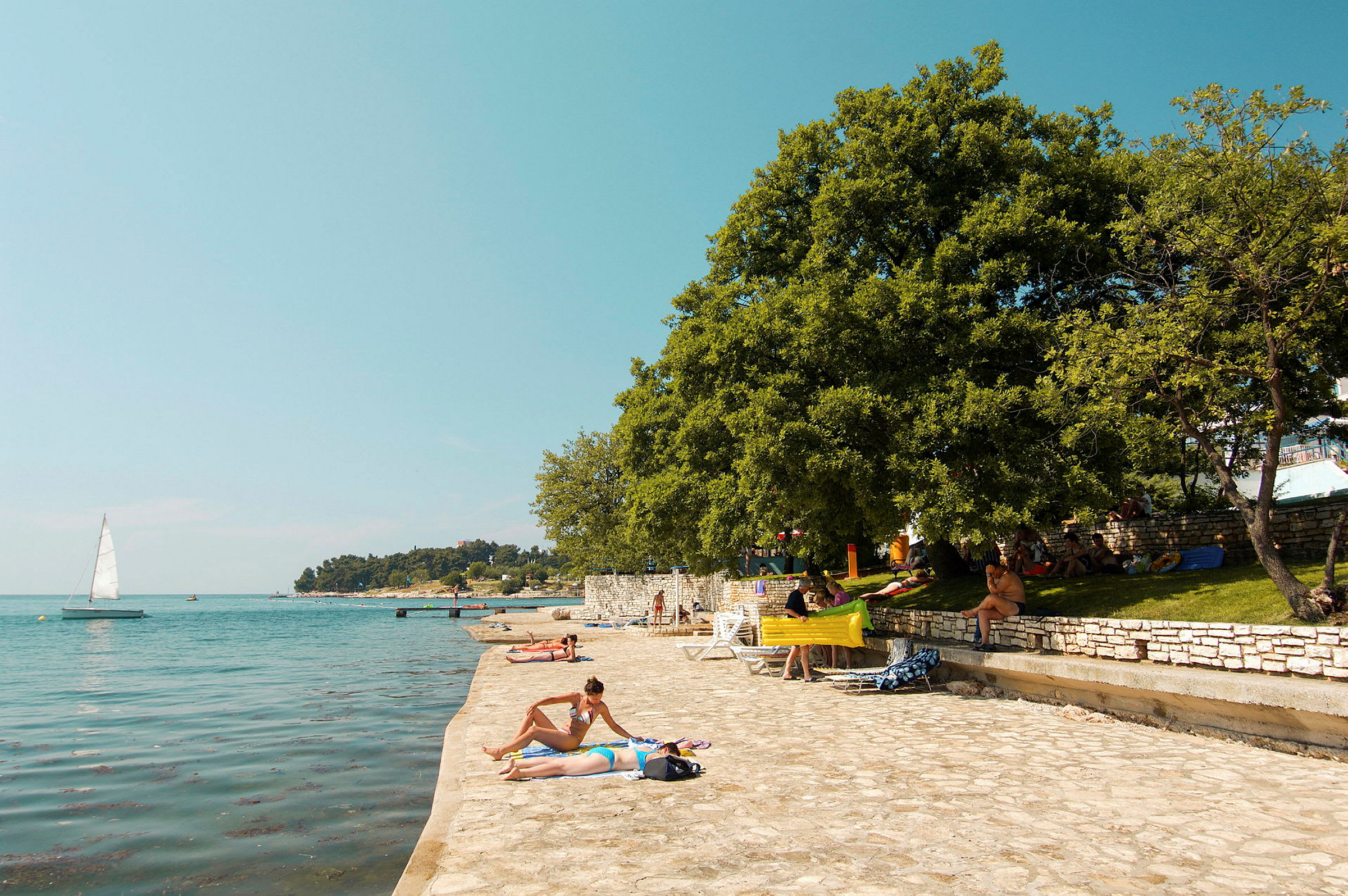 Personer som soler ved en steinsatt strand med trær i bakgrunnen og seilbåt på havet. (AI-generert tekst)