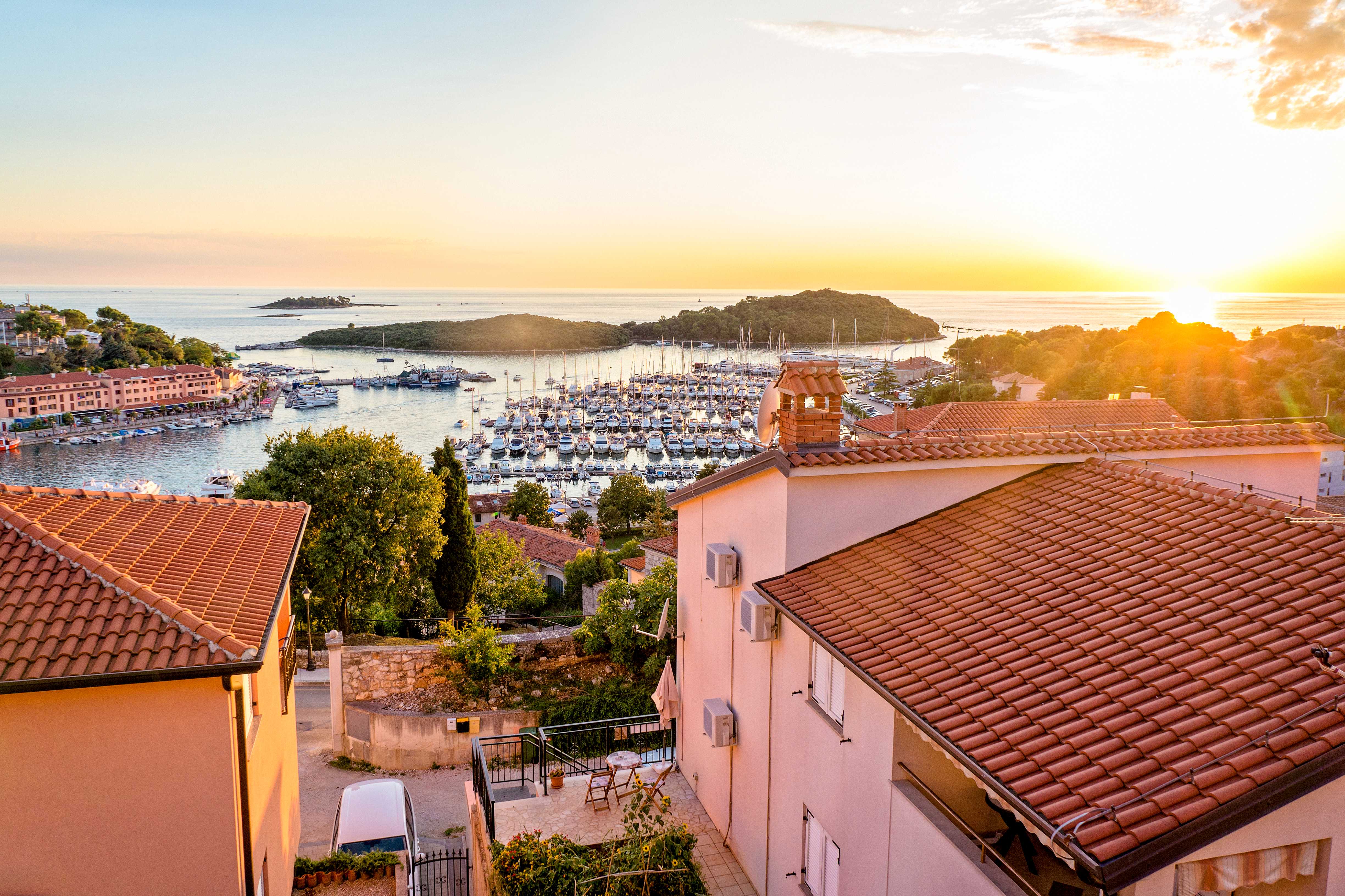 Utsikt over en småbåthavn med seilbåter omgitt av hus og grøntområder ved solnedgang. (AI-generert tekst)