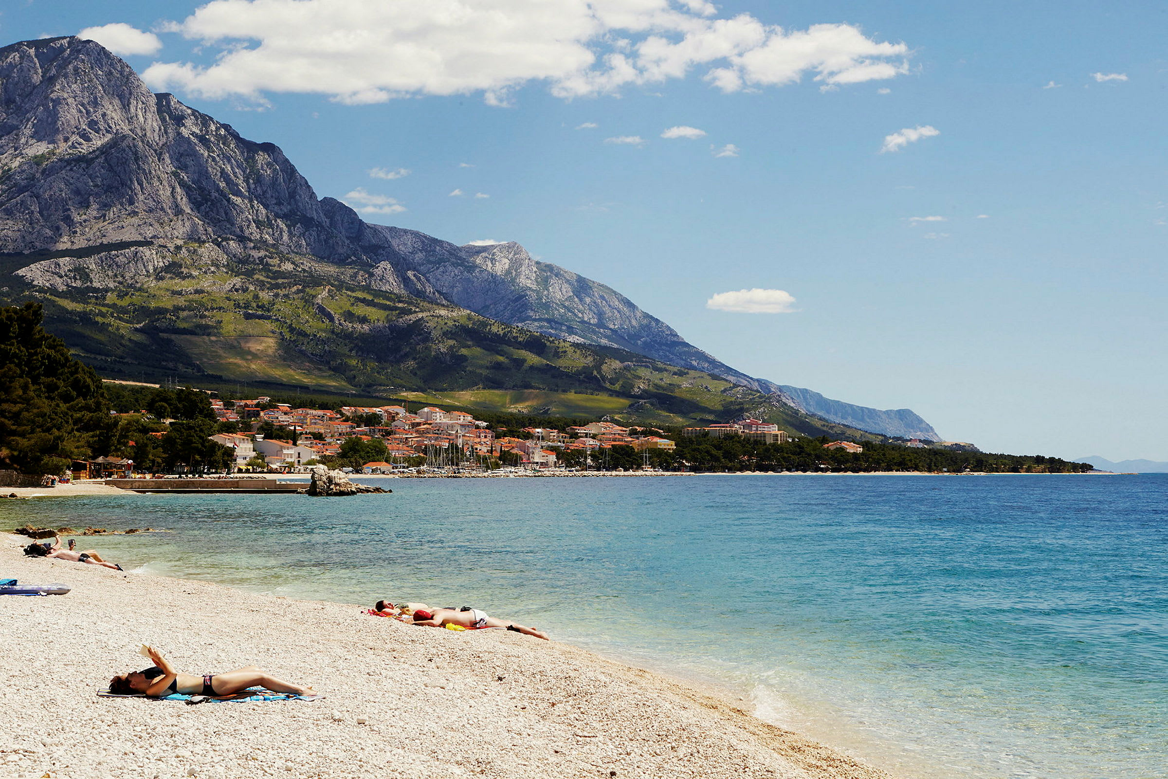 En strandscene med personer som soler seg, kystby i bakgrunnen og fjell. (AI-generert tekst)