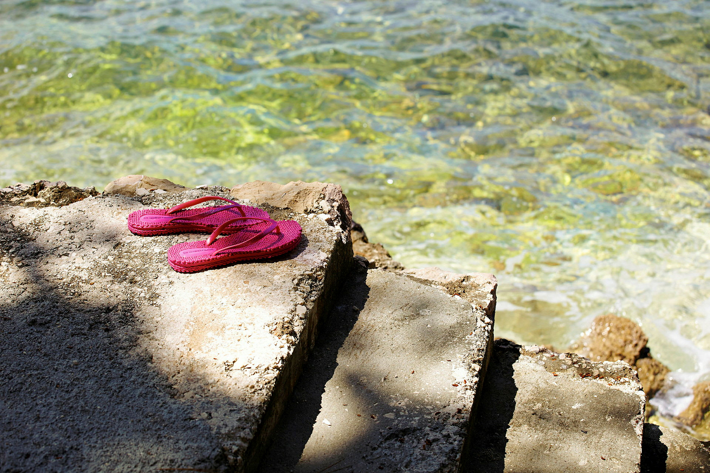 Rosa flip-flops ligger på en sementtrapp ved klart vann. (AI-generert tekst)