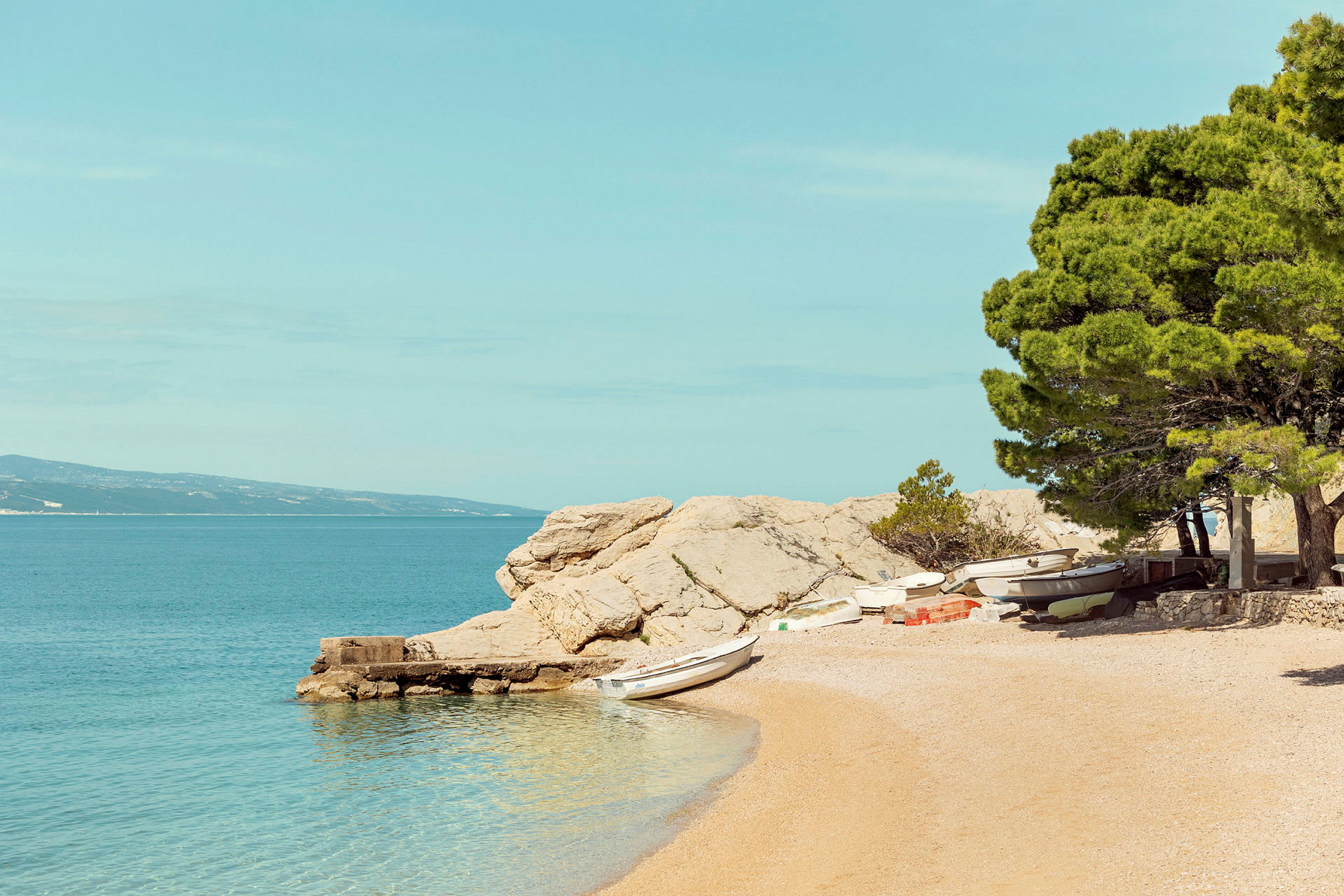 En strand med småbåter, trær og klipper ved vannet. (AI-generert tekst)