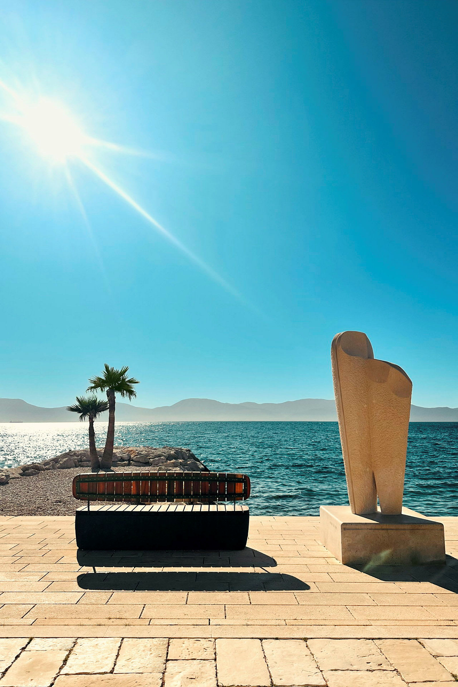 En strandpromenade med en solbenk og en skulptur, med havet og solen i bakgrunnen. (AI-generert tekst)