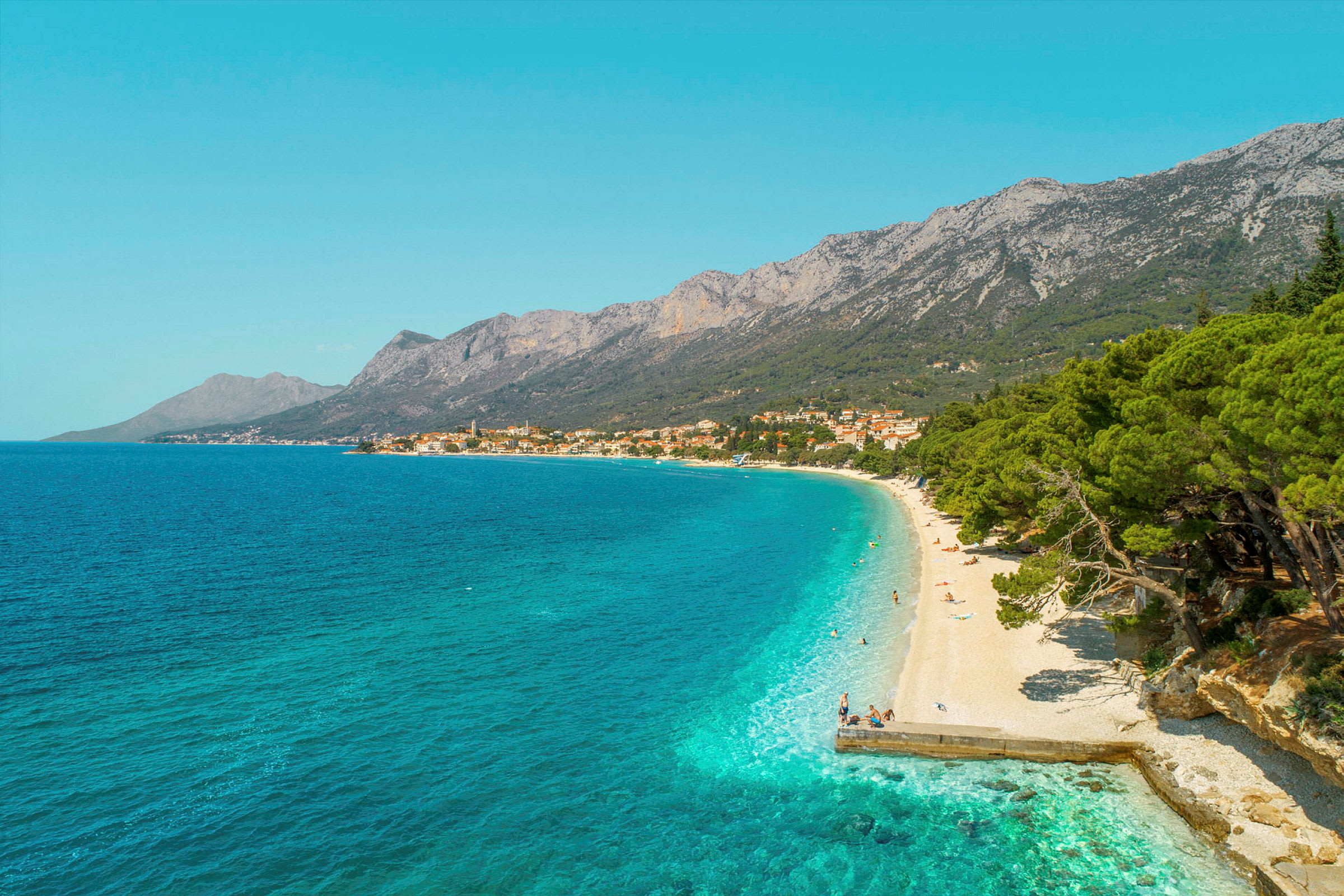 Utsikt over en rullesteinstrand med turkist vann og frodige trær. Fjell og en landsby vises i bakgrunnen. (AI-generert tekst)