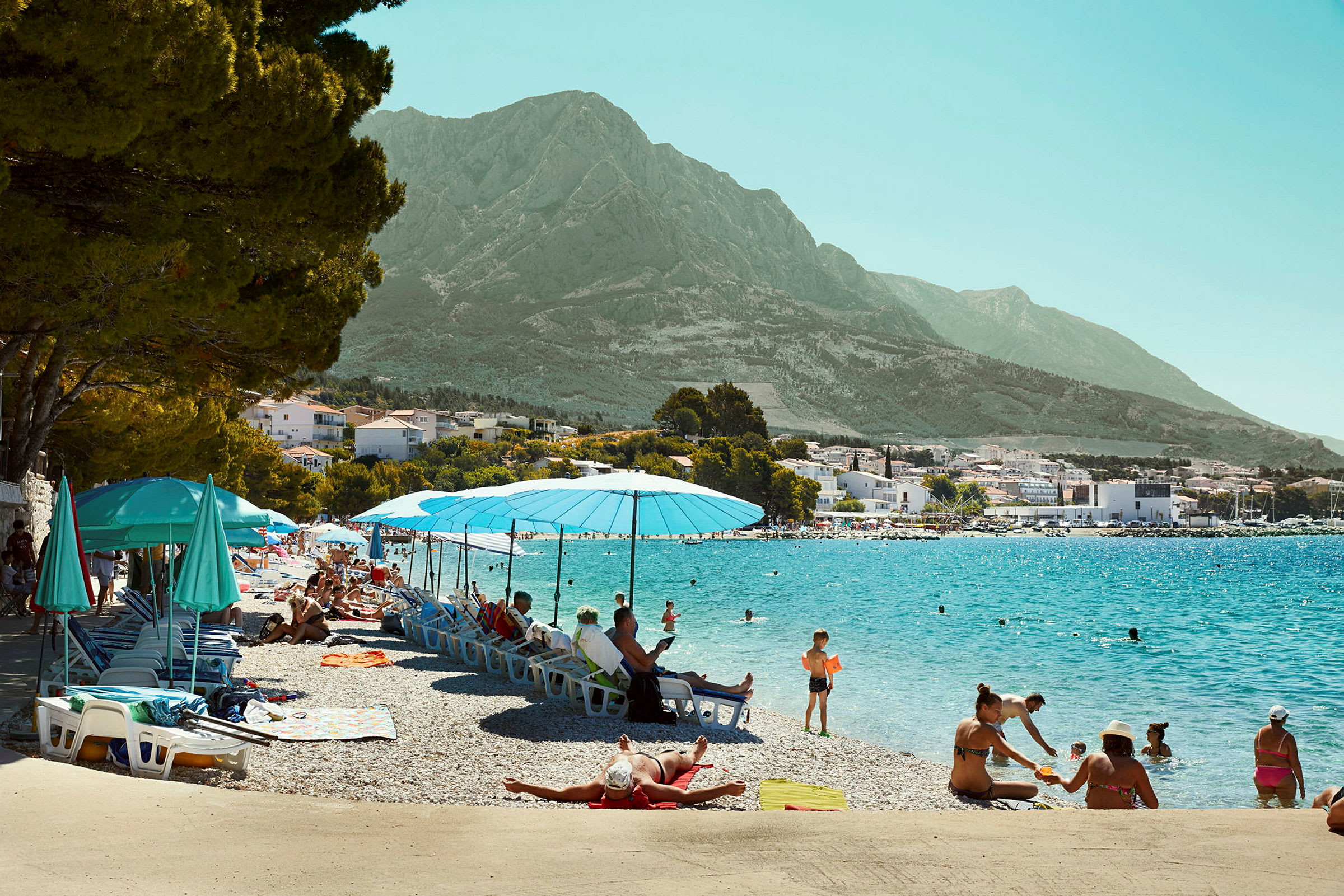Strand med parasoller og solsenger ved klart vann, omgitt av fjell. (AI-generert tekst)