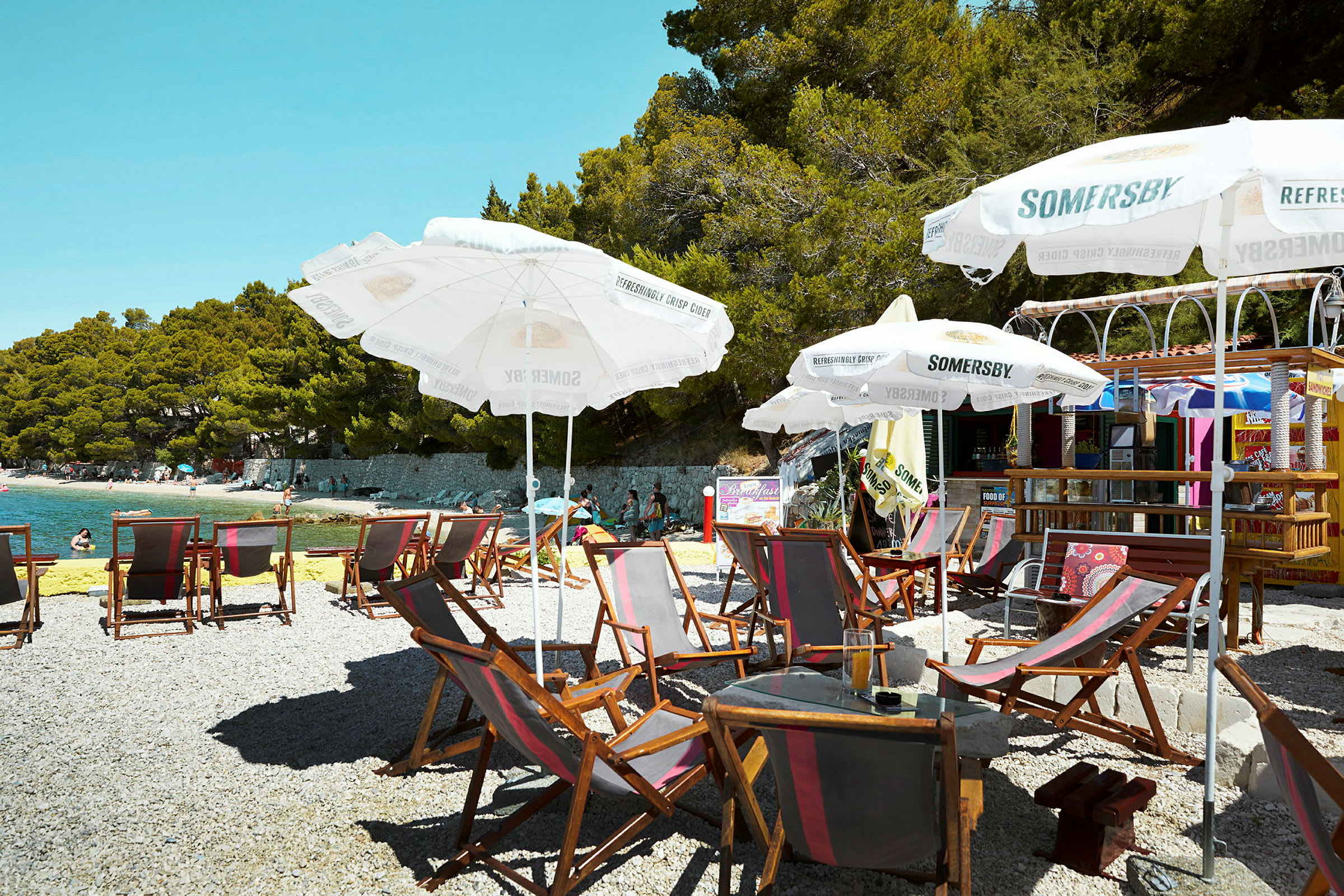 Strand med solsenger, parasoller og en kafé i bakgrunnen. (AI-generert tekst)