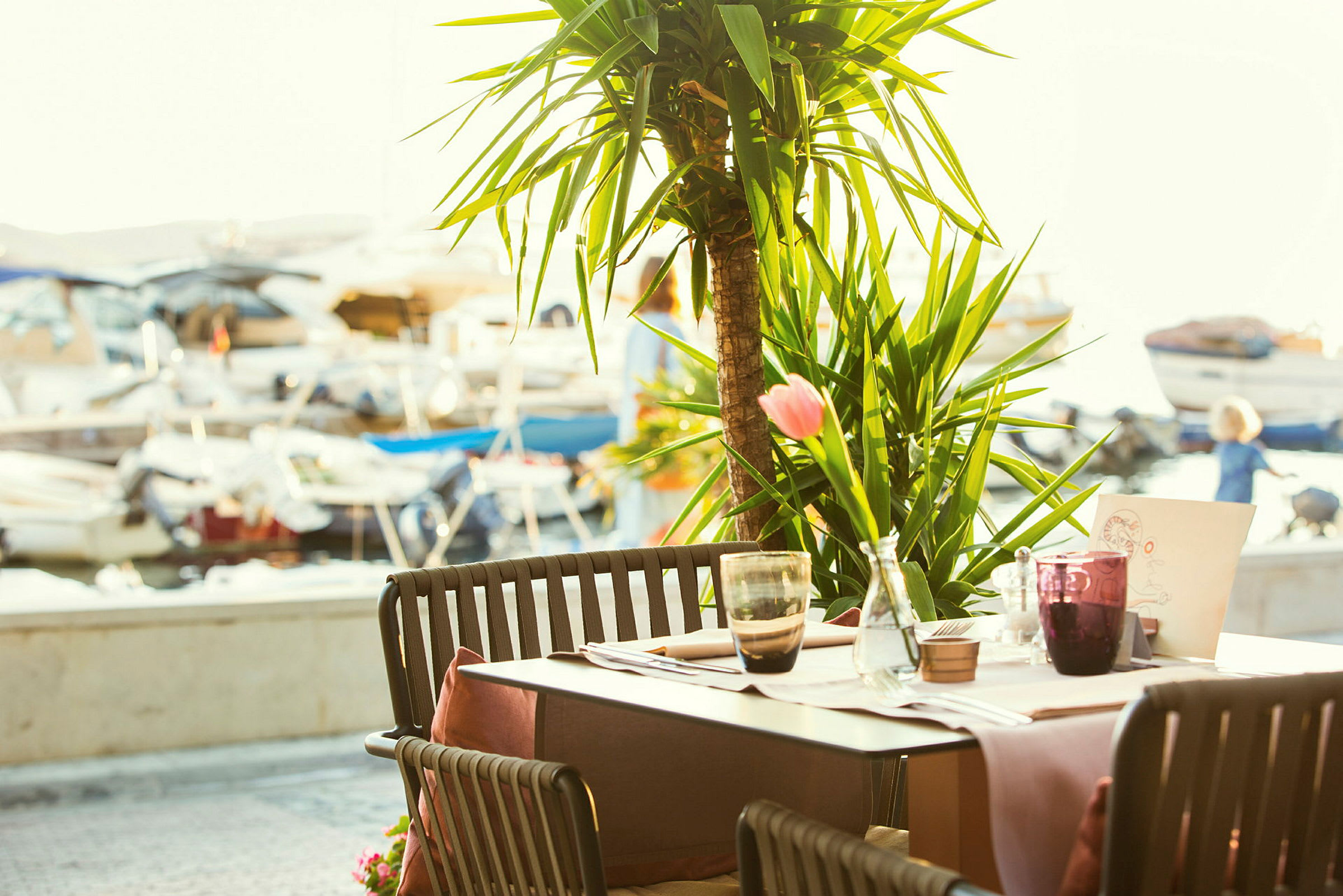 En uterestaurant med bord og stoler med en palme i bakgrunnen og utsikt over småbåter ved en brygge. (AI-generert tekst)