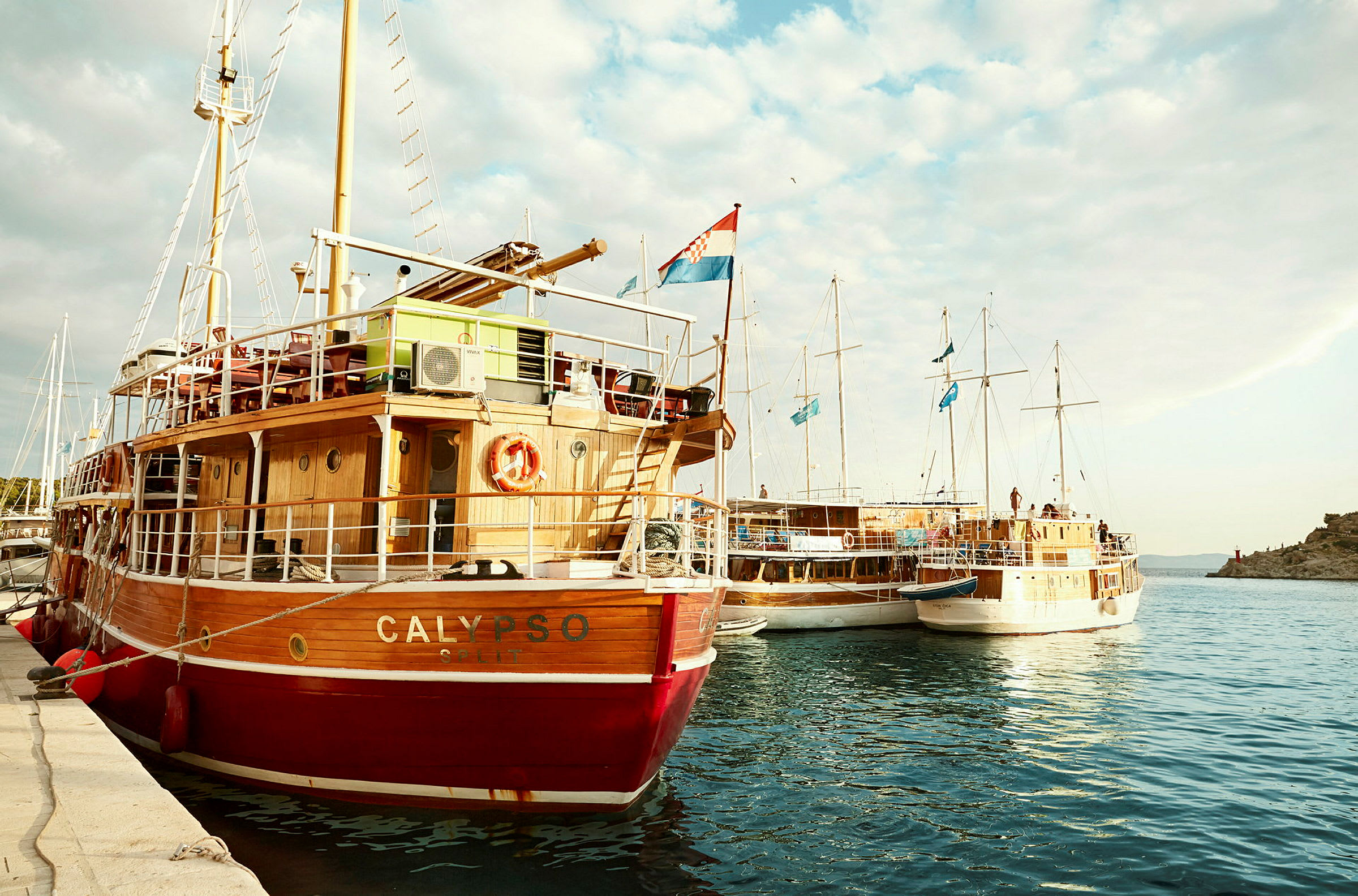 En rød og hvit seilbåt ved navn Calypso ved en brygge, omgitt av andre båter med blå flagg. (AI-generert tekst)