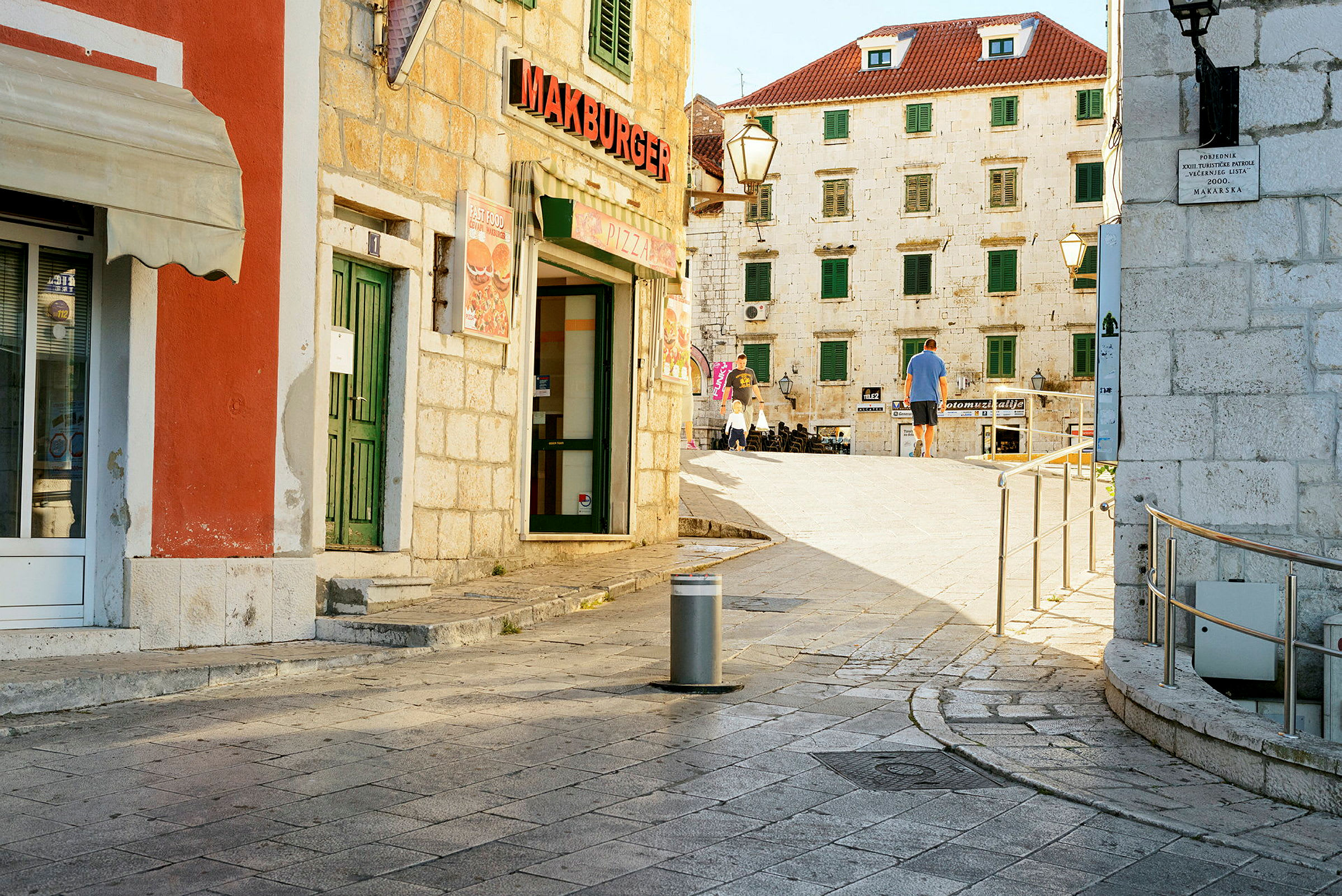 Bymiljø med steinbelagt gate og bygninger med røde tak og grønne skodder, samt en restaurantskilt med teksten 'Makburger'. (AI-generert tekst)