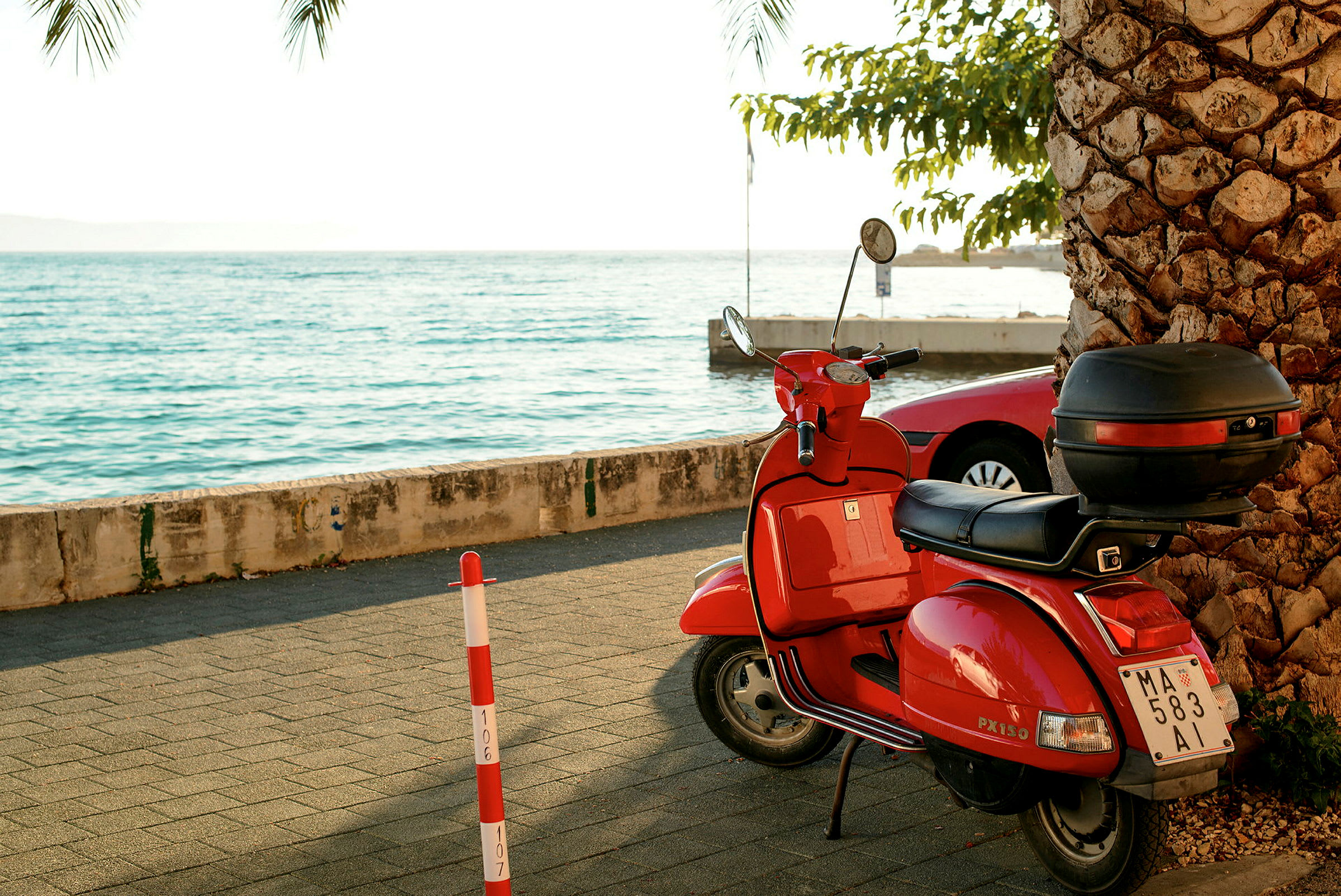Rød moped parkert på gangvei nær sjøen, omgitt av en palme og parkerte biler. (AI-generert tekst)