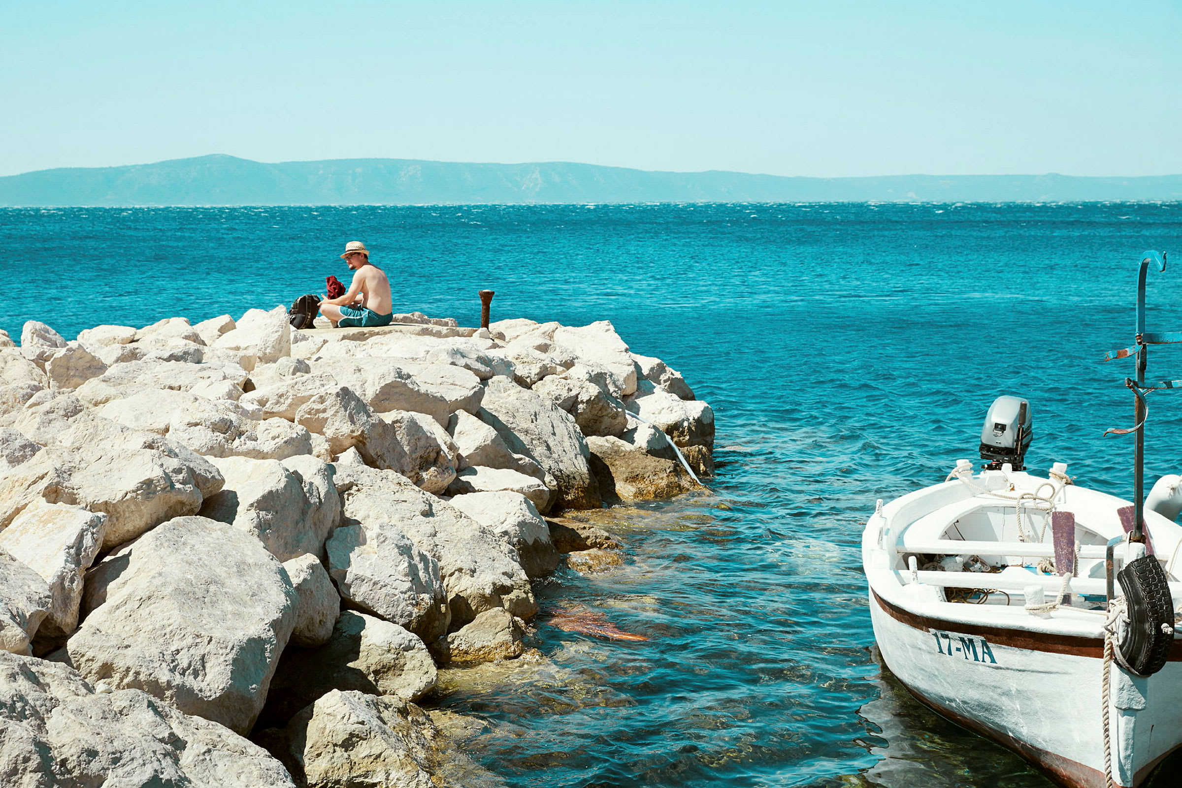 En person sitter på steiner nær havet, med en liten båt fortøyd ved siden av. (AI-generert tekst)