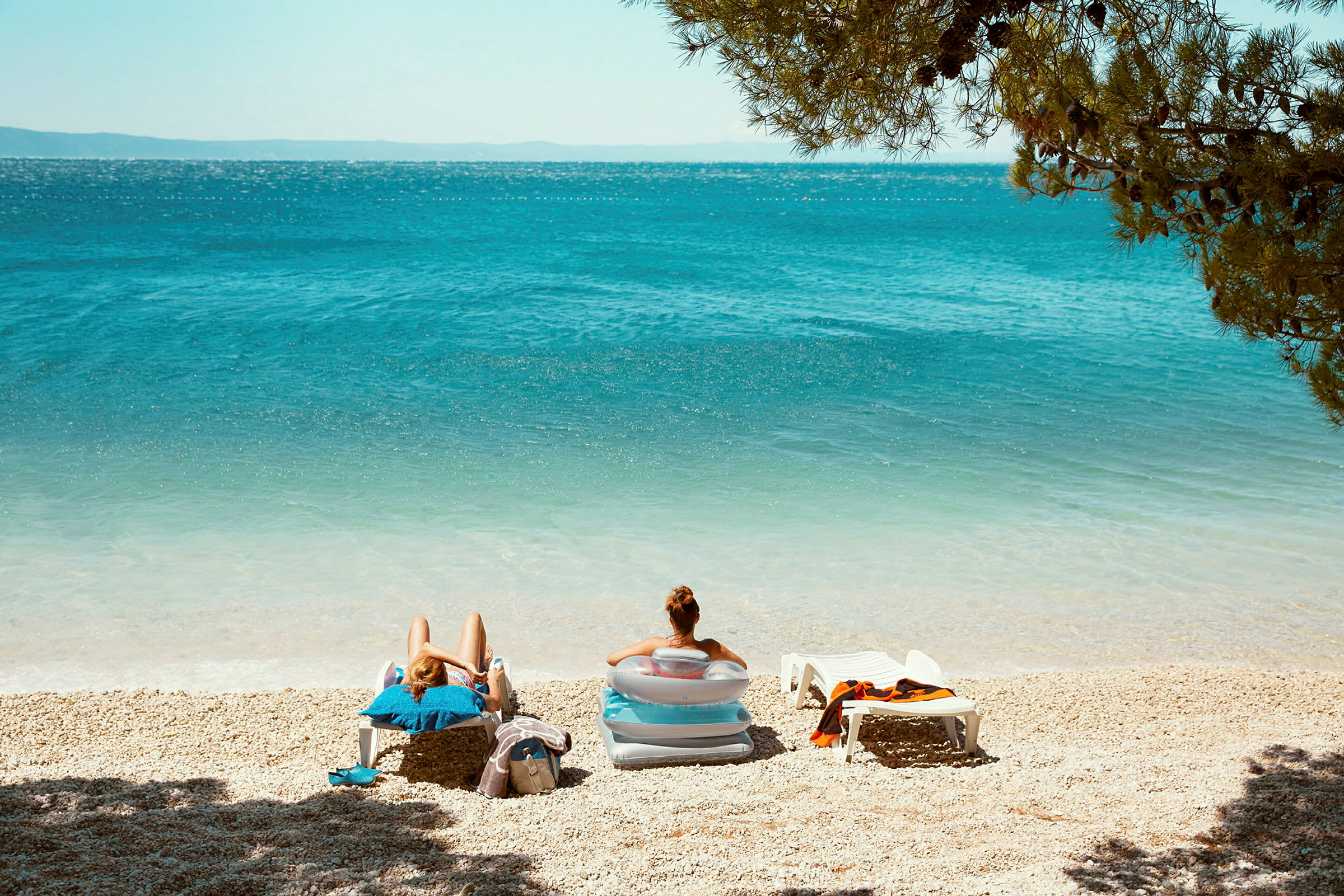 To personer sitter i solsenger på en strand med blått hav og omkringliggende grøntområder. (AI-generert tekst)
